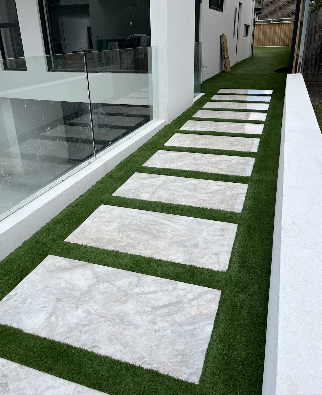 A modern outdoor pathway with rectangular white stone tiles set in green artificial grass, alongside a building with large glass windows.