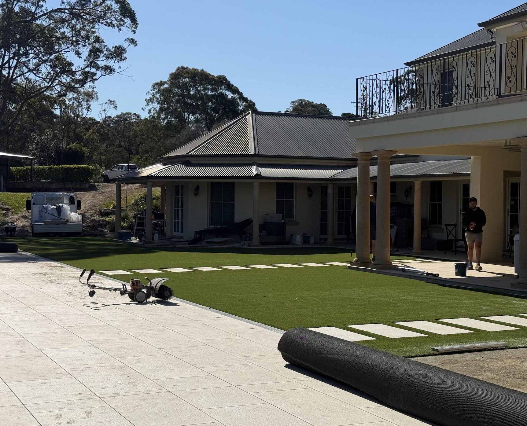 Construction workers installing artificial grass in a backyard area of a house, with tools and equipment on the ground, and a house with a patio and balcony in the background.