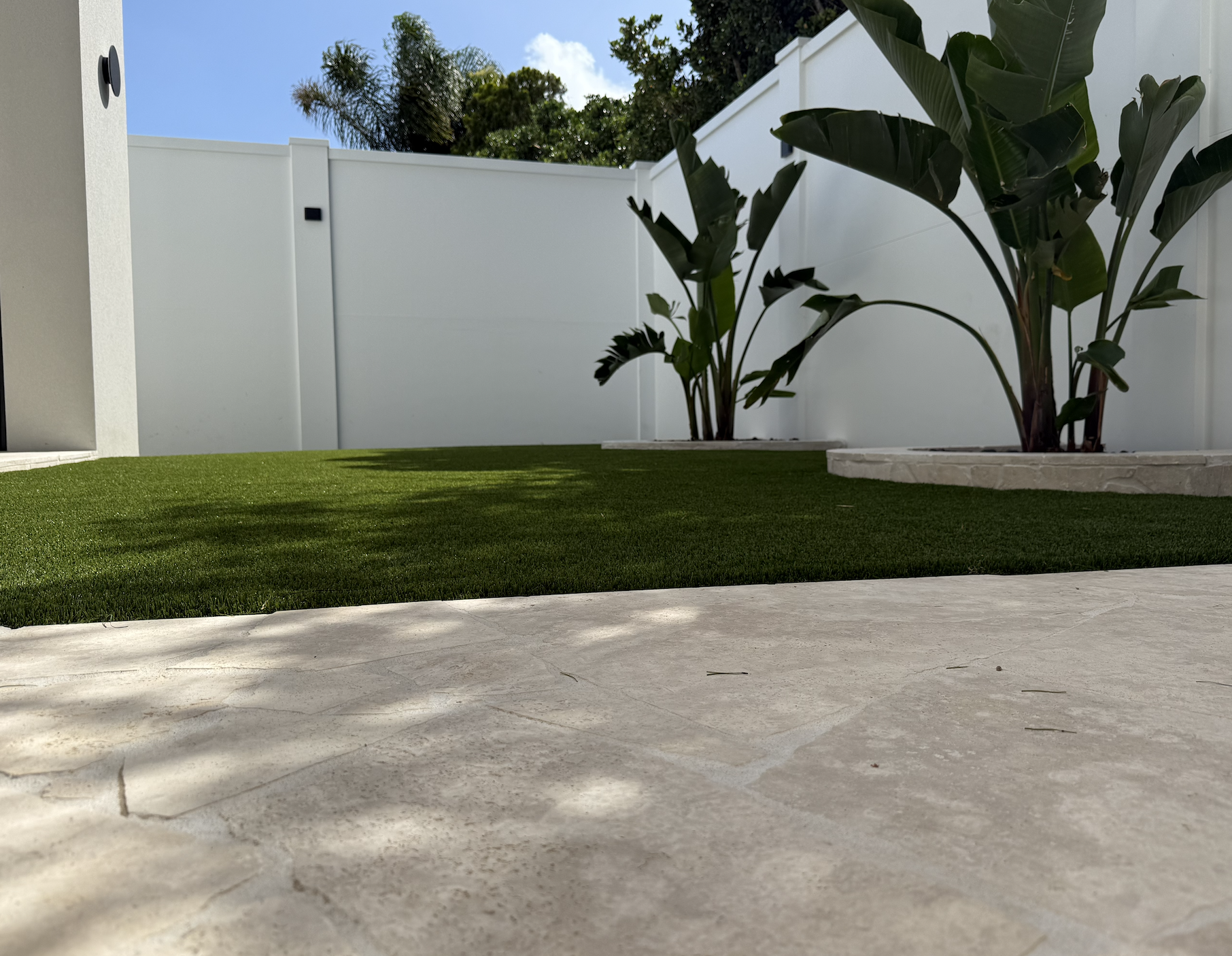 A backyard with a small patch of artificial grass, a concrete patio, tall green plants in white planters, a white privacy fence, trees, and a blue sky with some clouds.