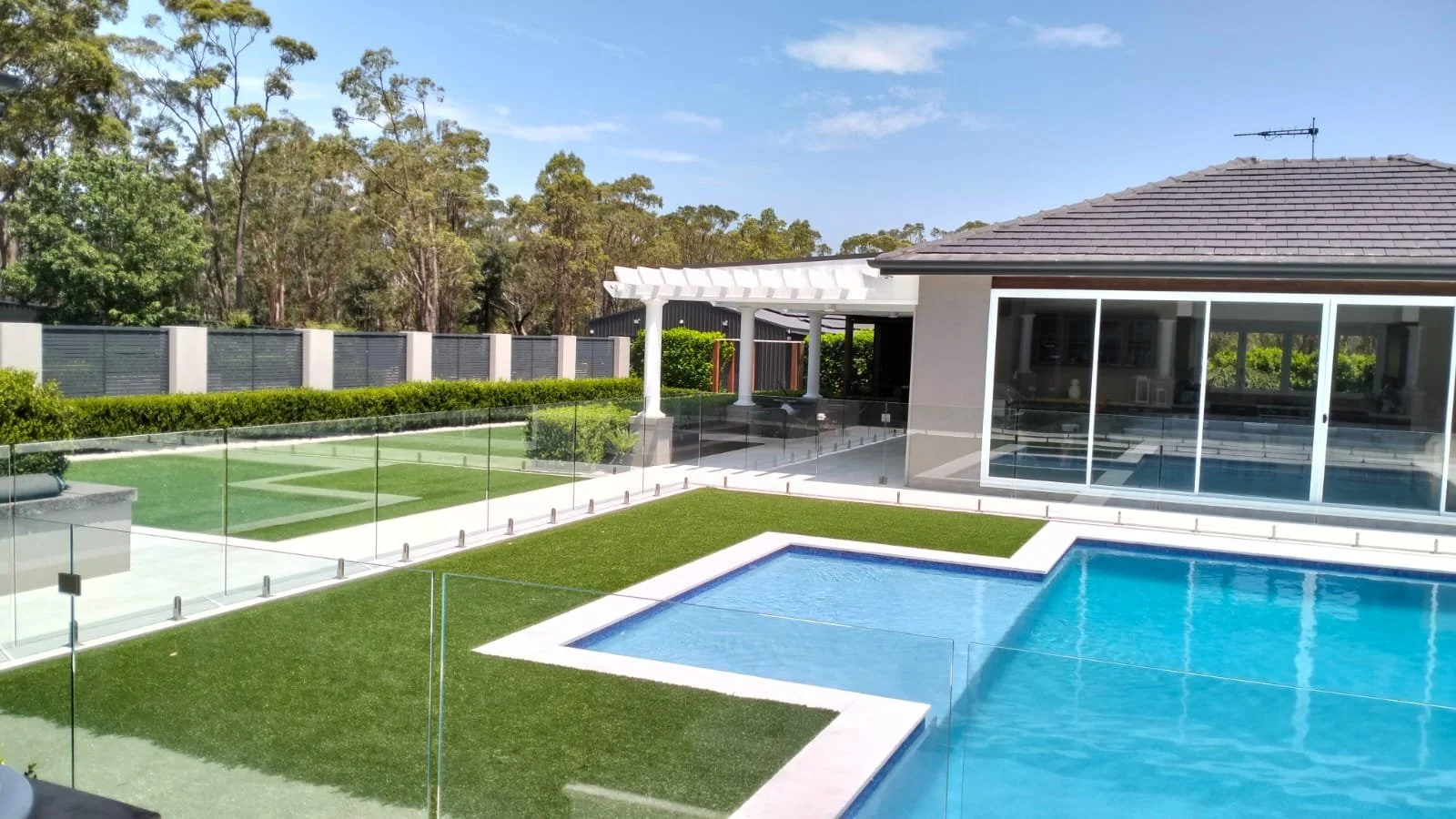 A backyard with a swimming pool, a separate hot tub, a glass fence, a patio with white pergola, and a house with large sliding glass doors; surrounded by green bushes and trees.