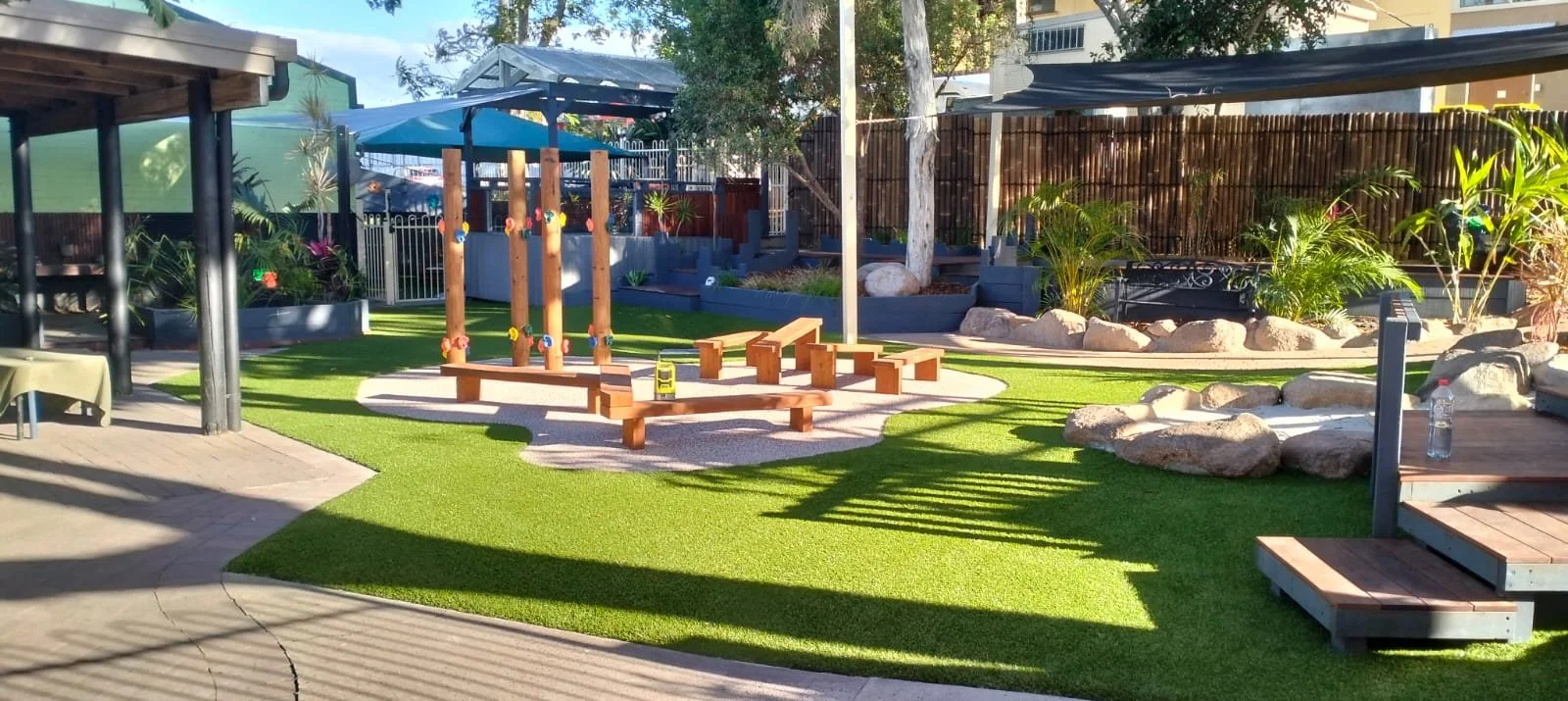 A backyard playground area with artificial grass, a wooden climbing structure with ropes, benches, rocks, surrounded by plants and trees, shaded areas, and outdoor furniture.