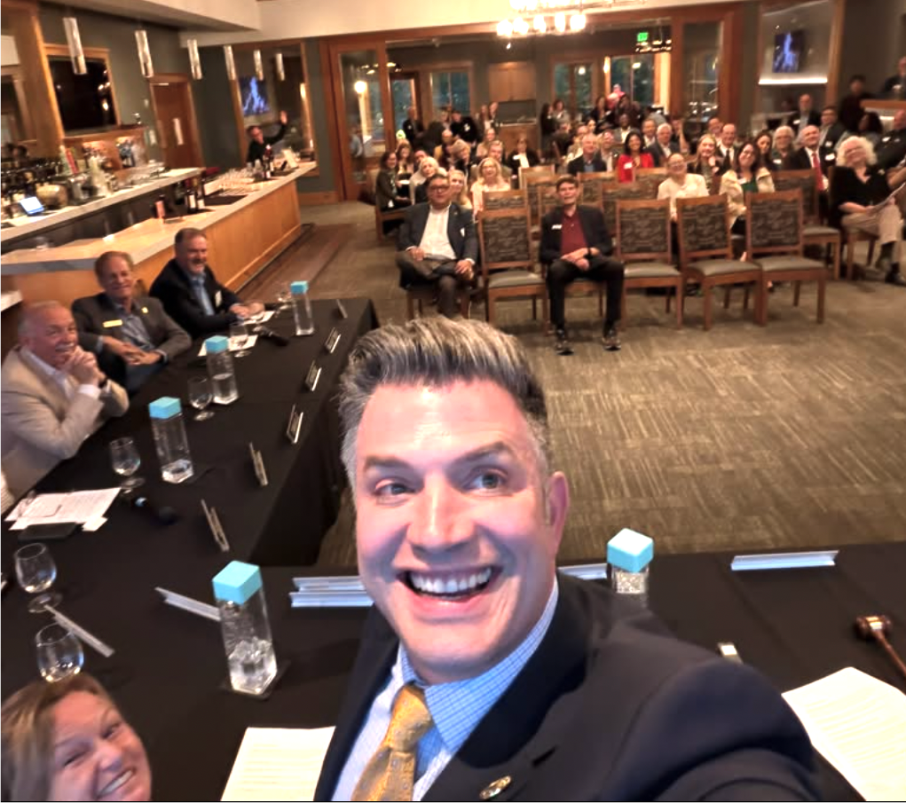 A smiling man in a suit taking a selfie at a conference or event, with a large audience seated behind him in a well-lit room.
