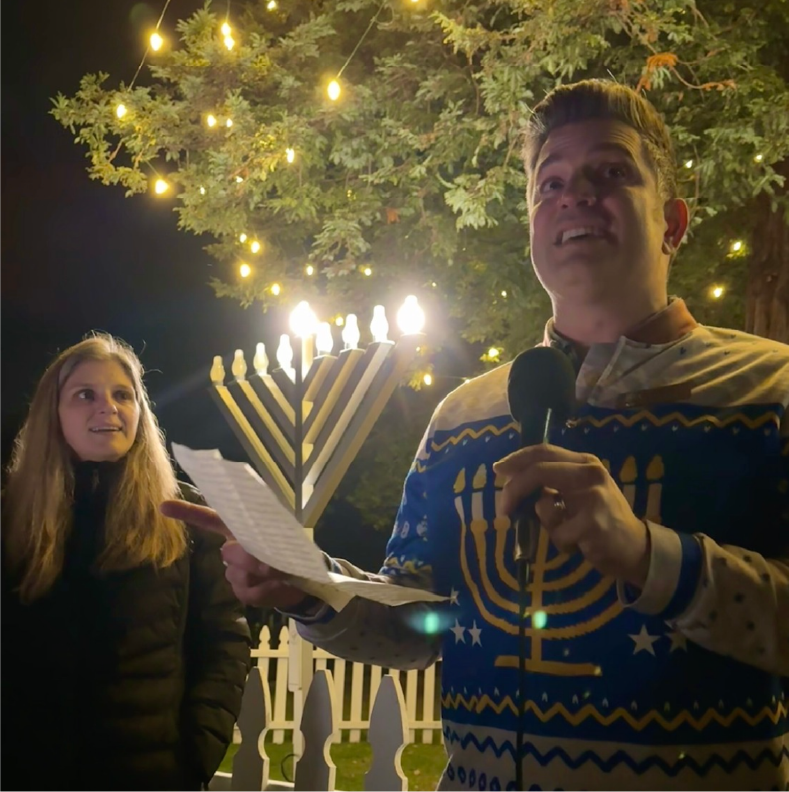 A man holding a microphone and reading from a paper in front of a menorah, with a woman watching, decorated with string lights, in an outdoor setting at night.