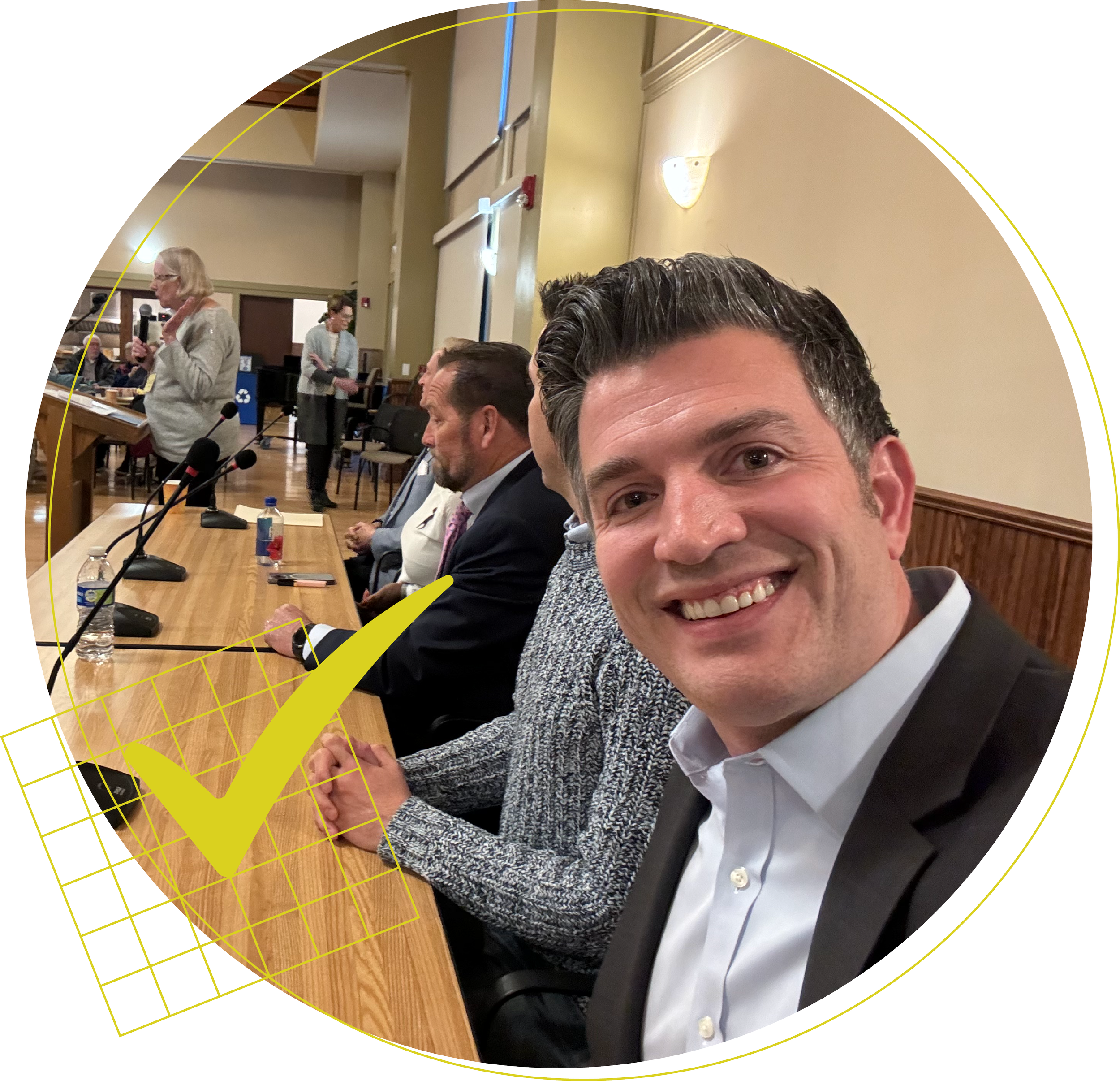 Kerry Hillis, candidate for re-election to Moraga Town Council, smiling at the camera during a meeting with a panel of people, women and men, seated at a conference table with microphones and papers in a room with beige walls and wood paneling.