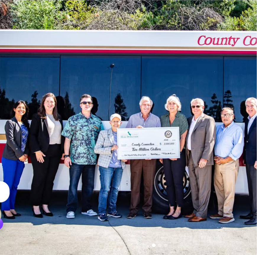 Group of nine people, standing outdoors in front of a bus, holding a large ceremonial check for two million dollars in County Connection funds, with trees behind them.