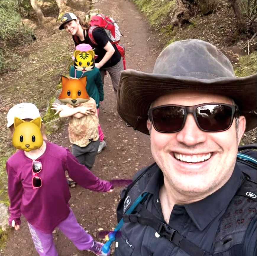 A group of five people, including two children, hiking on a dirt trail in a forested area. The person in the foreground is a man wearing a wide-brimmed hat, sunglasses, and a black shirt, smiling at the camera. The children and the woman behind him have animal face emojis over their faces for privacy.