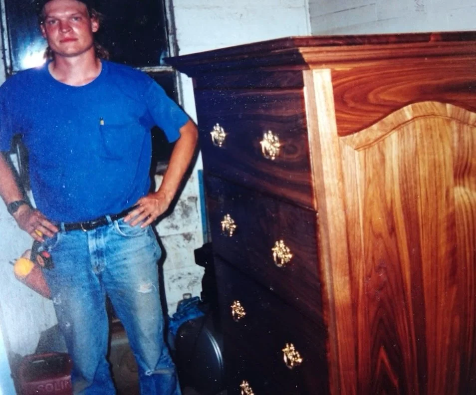A man standing next to a large wooden dresser in a room with a distressed wall. The man is wearing a blue shirt and jeans, holding tools in his right pocket, with a tool belt on his waist.