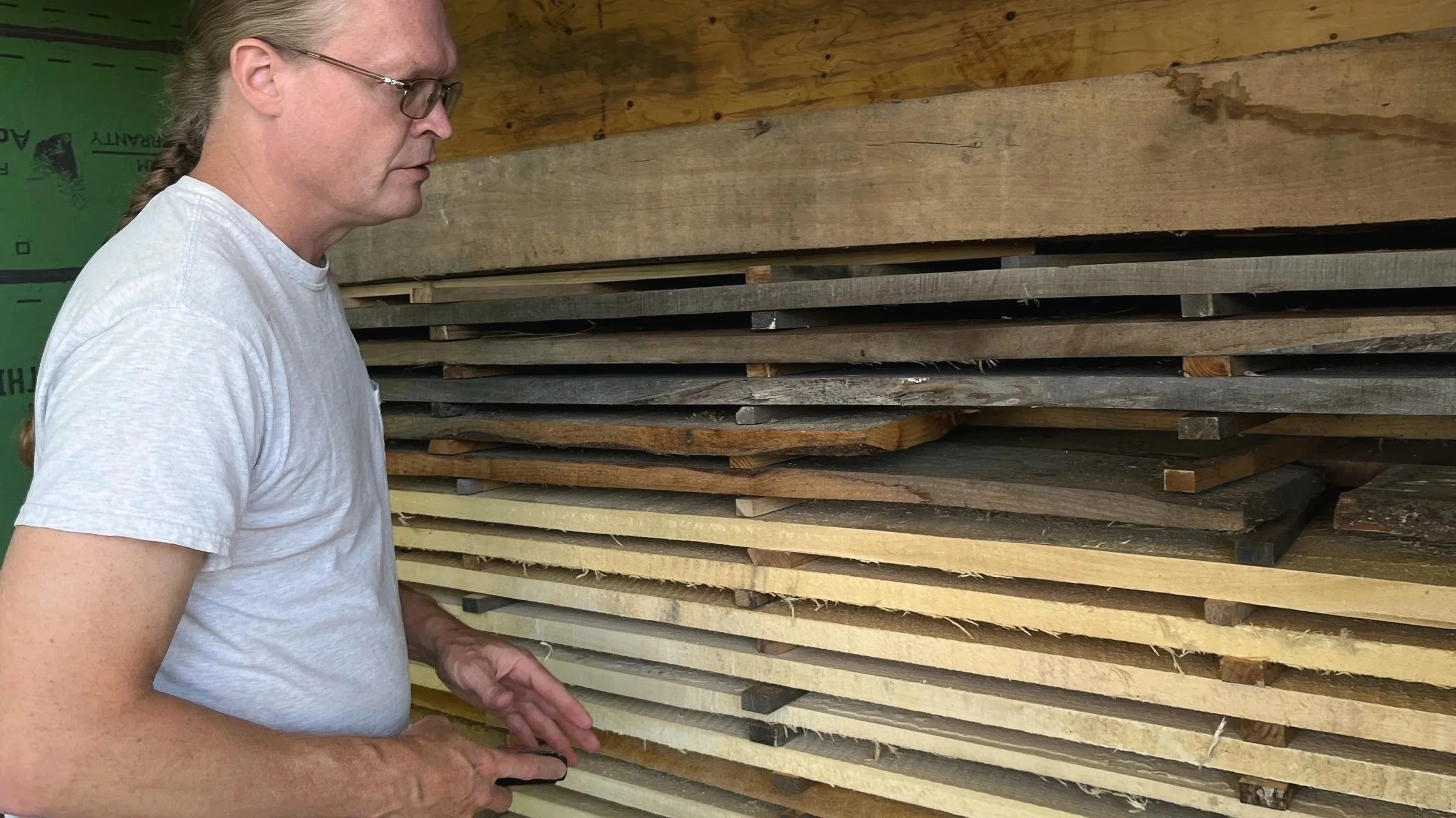 A person with glasses and long braided hair wearing a white T-shirt looking at stacked pieces of wood in a workshop.
