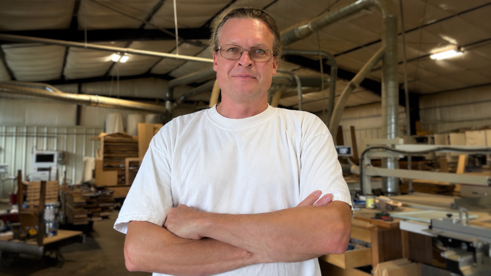 A man with glasses stands with arms crossed in a woodworking shop, surrounded by woodworking tools and stacks of wood.