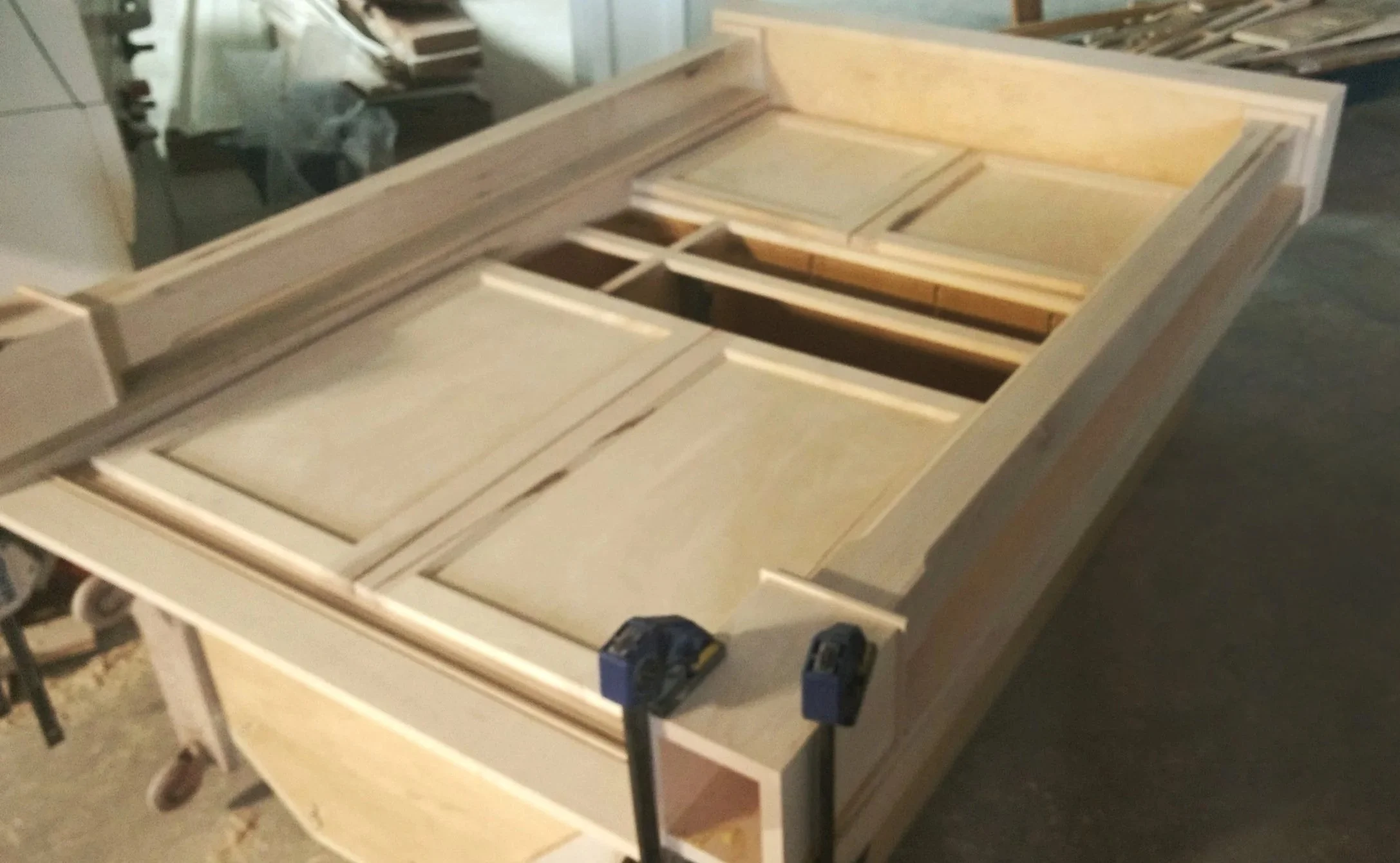 Unfinished wooden cabinet or shelving unit in a workshop, with clamps attached and tools nearby.