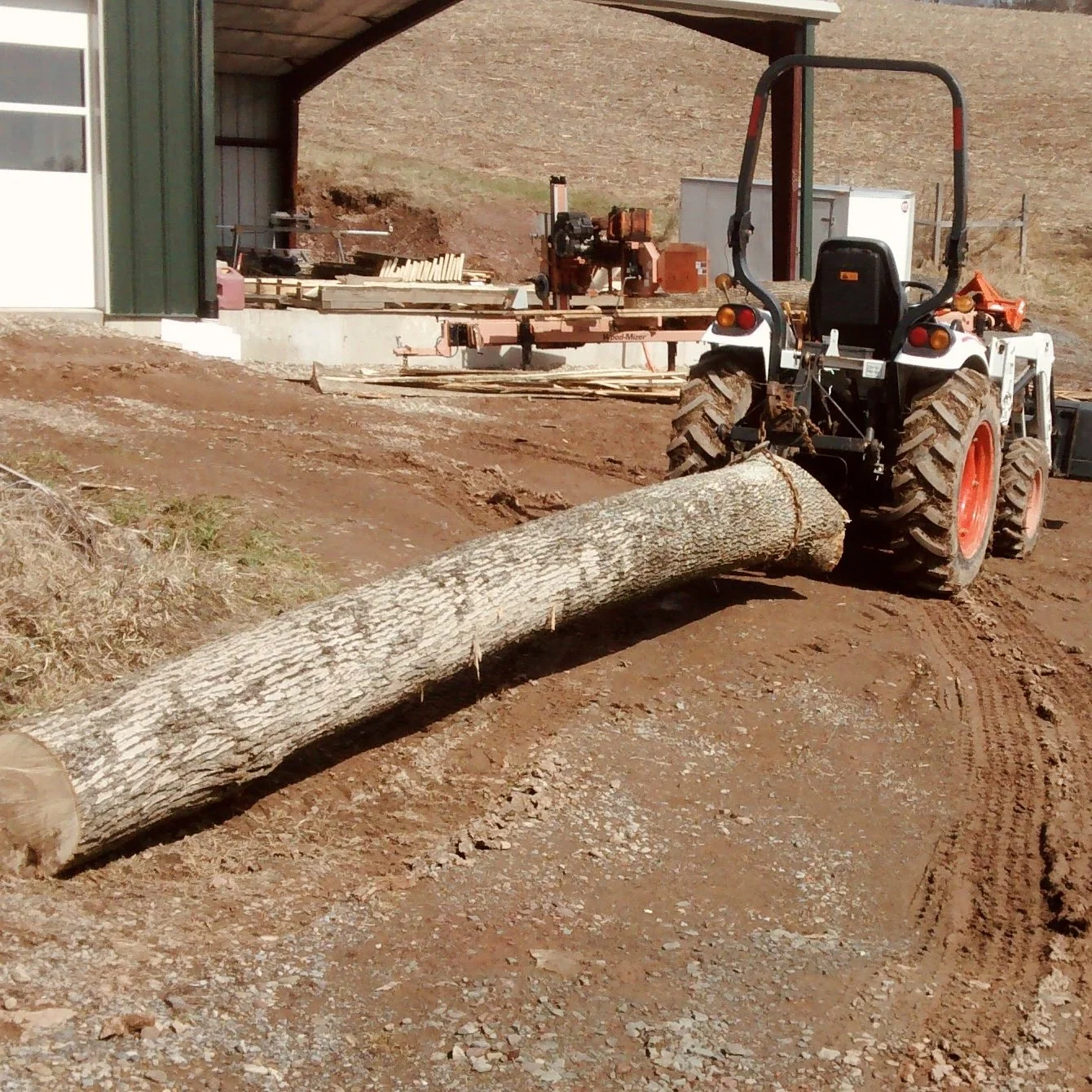 A tractor with large tires lifting a fallen tree log on a dirt ground near a building with construction tools and wood materials in the background.