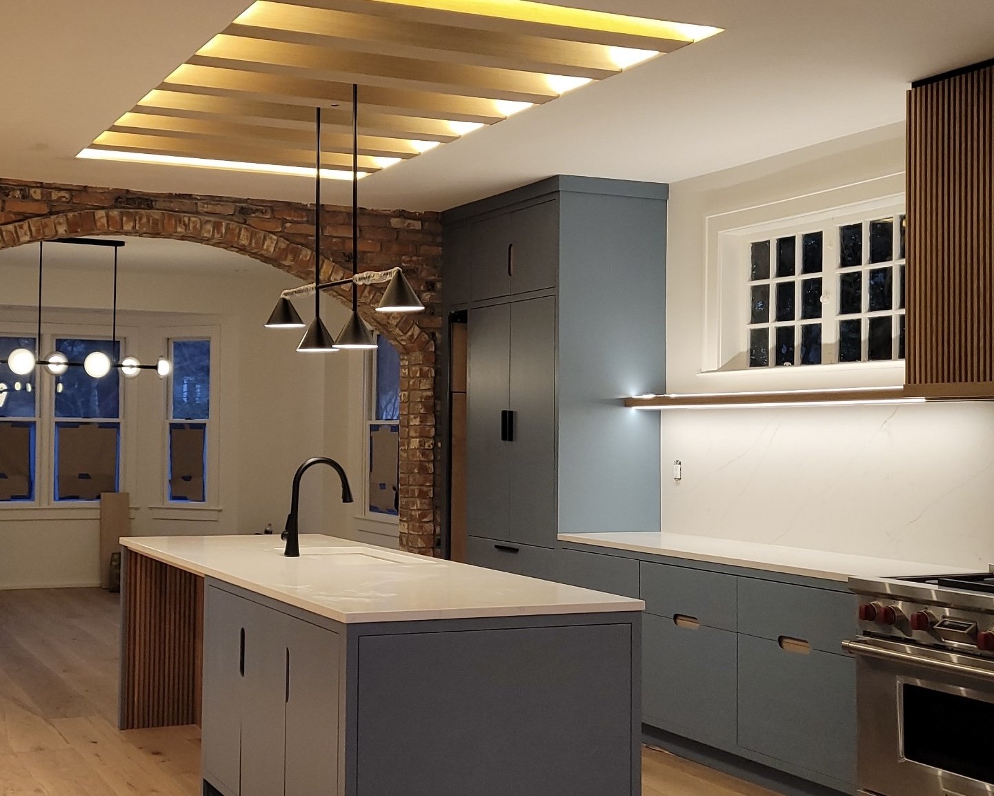 Modern kitchen with blue cabinets, white countertop, custom kitchen island, wood accents, brick archway, pendant lights, window with divided panes, and recessed lighting.