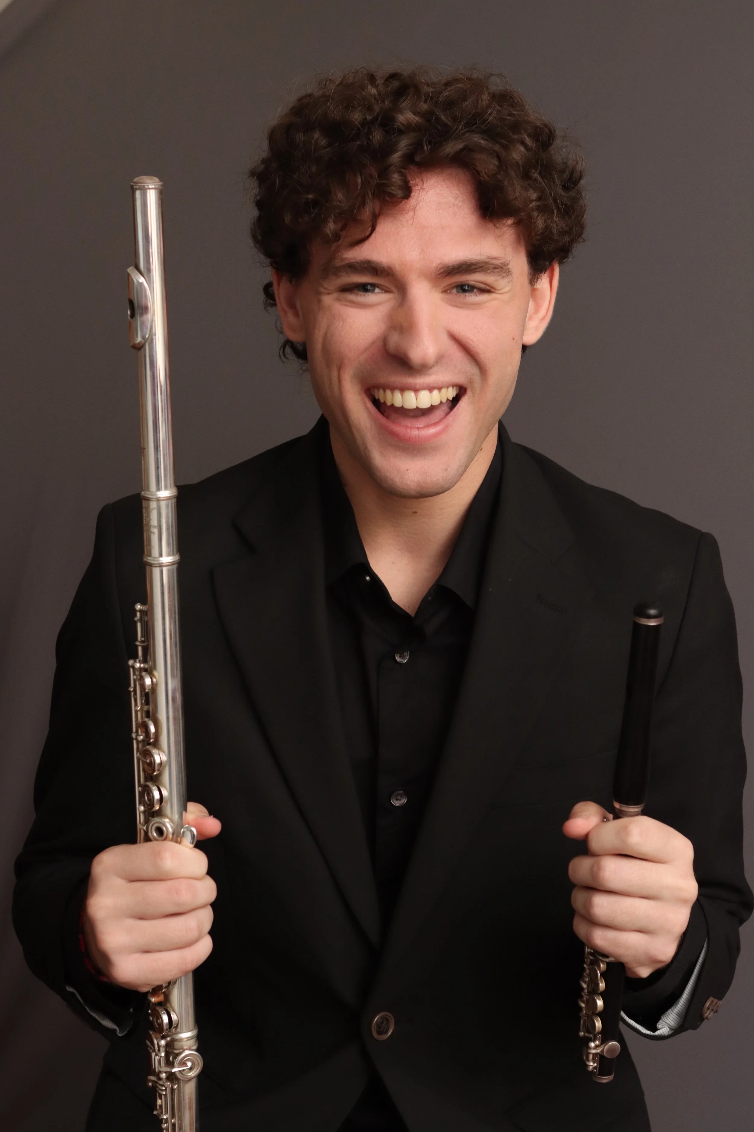 A smiling young man with curly brown hair dressed in a black suit, holding a silver flute in his right hand and a black and silver piccolo in his left hand, standing against a dark gray background.