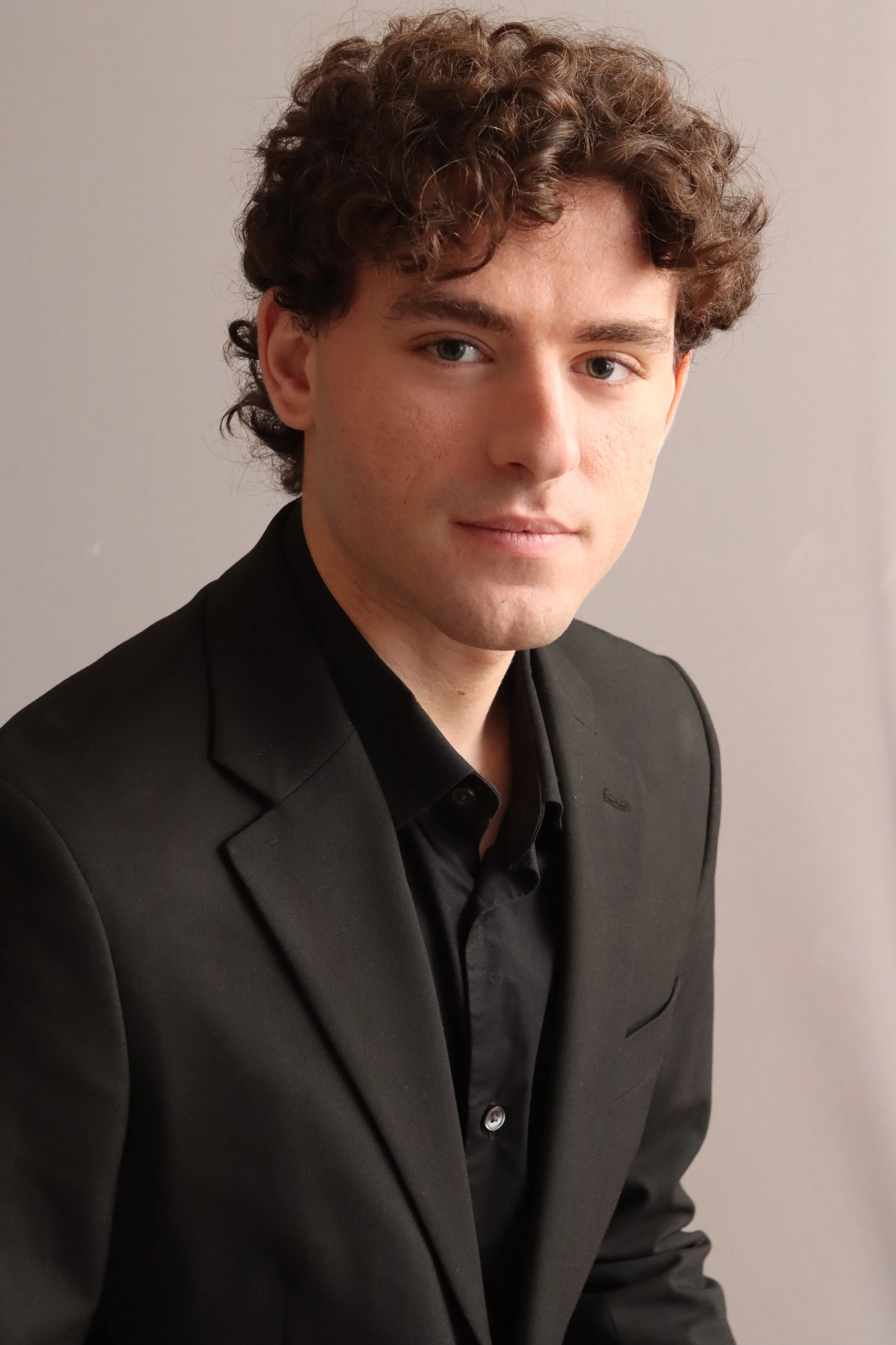 A young man with curly brown hair and light skin wearing a black suit and black shirt, posing against a plain light-colored wall.