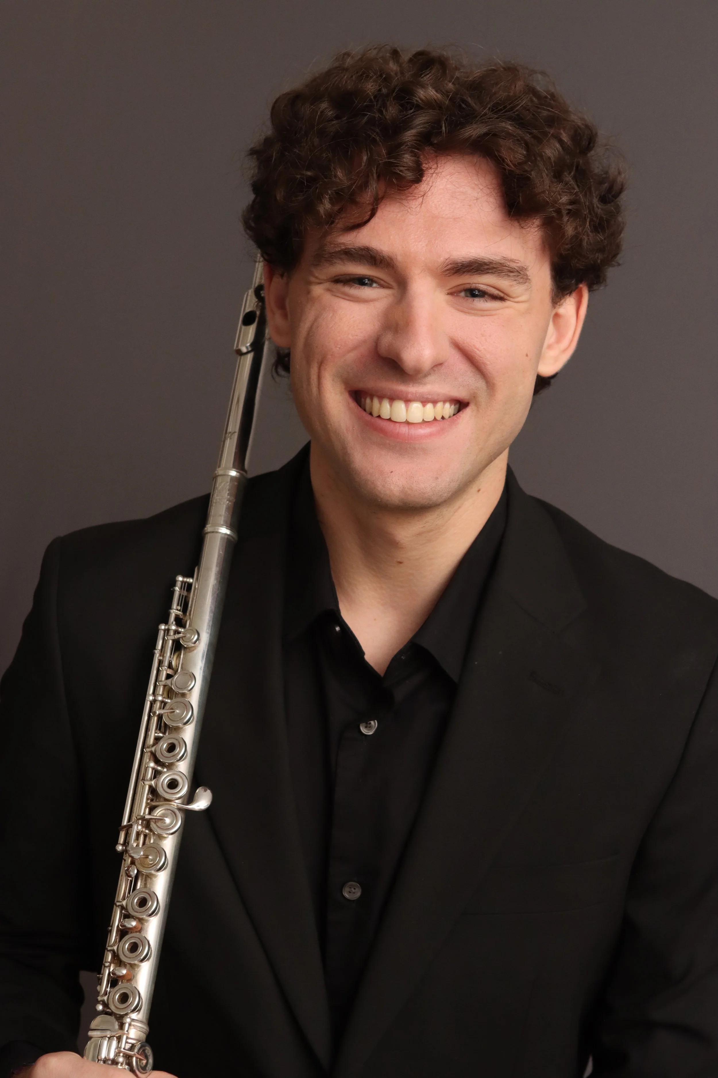 A man wearing a black suit holding a silver flute and smiling, against a plain dark background.