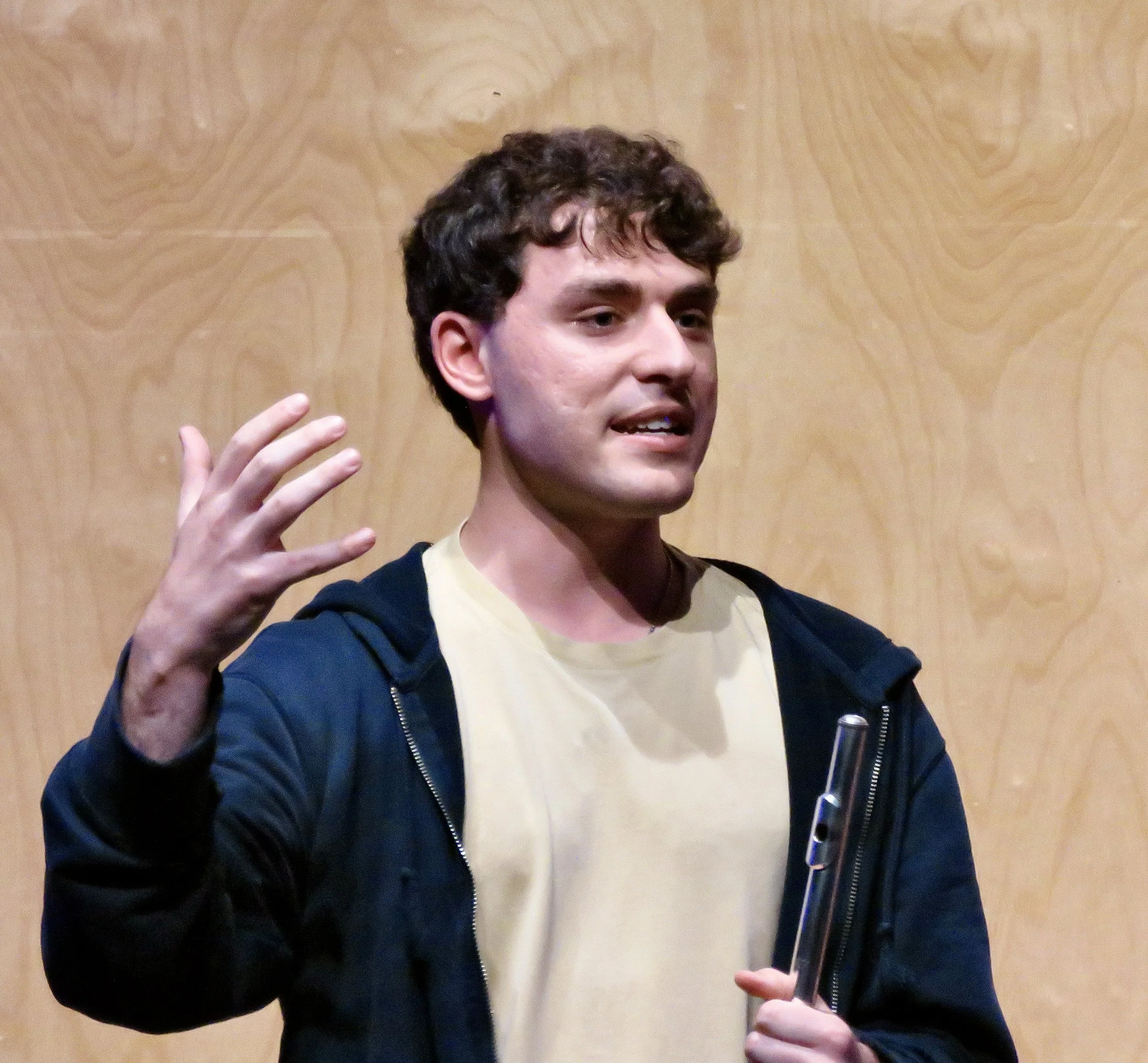 Young man with curly dark hair speaking or gesturing with his hands, wearing a black hoodie and a light-colored shirt, standing against a wooden textured background.