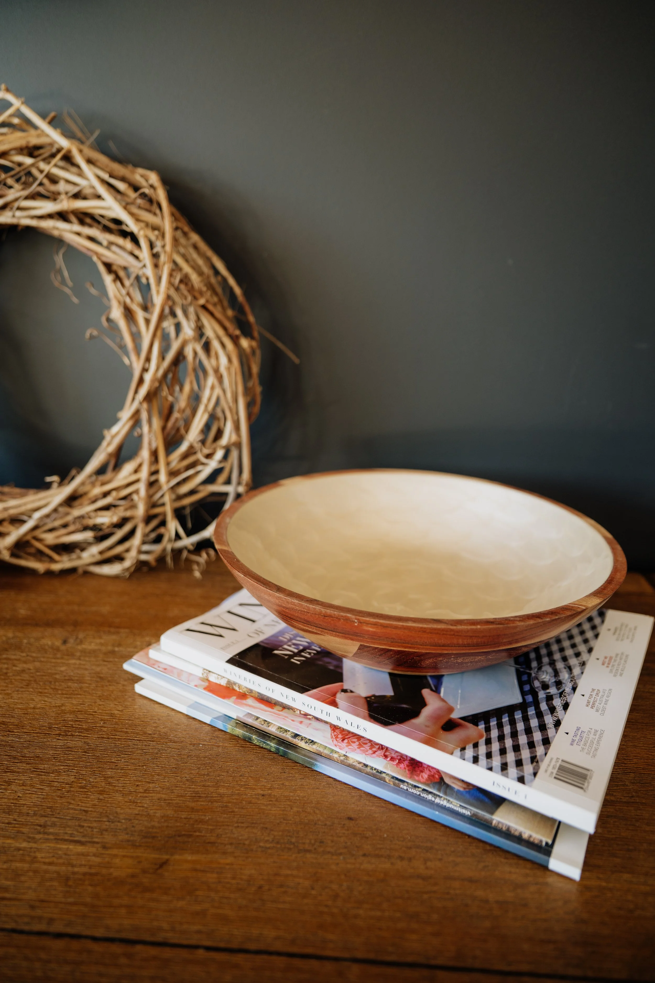 A woven wreath, an empty bowl, and a stack of magazines on a wooden table against a dark wall.