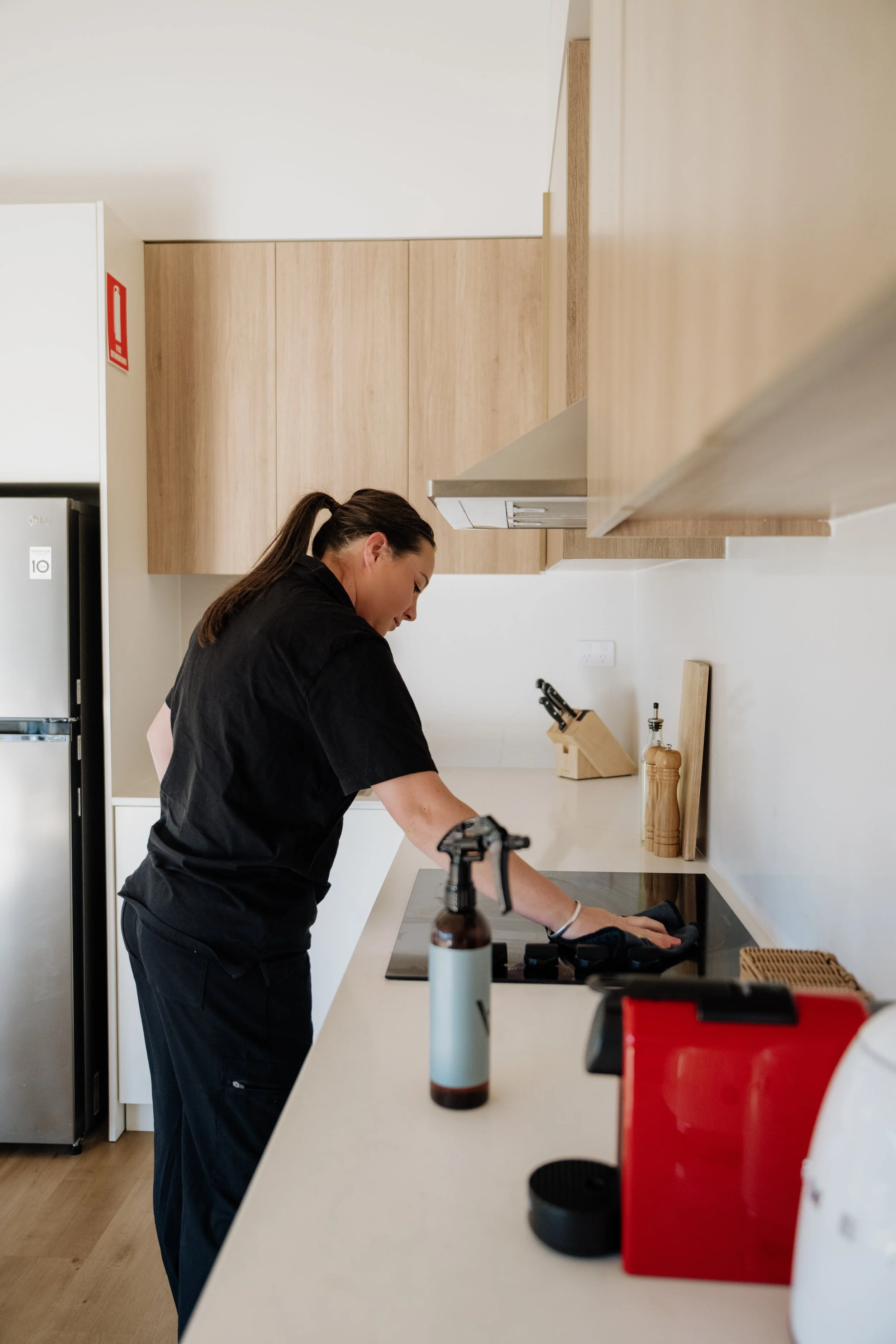 A woman with a ponytail and wearing a black shirt is cleaning a kitchen countertop with a cloth. The kitchen has wooden cabinets, a stove, and various items like a spray bottle, a red appliance, and a utensil holder.