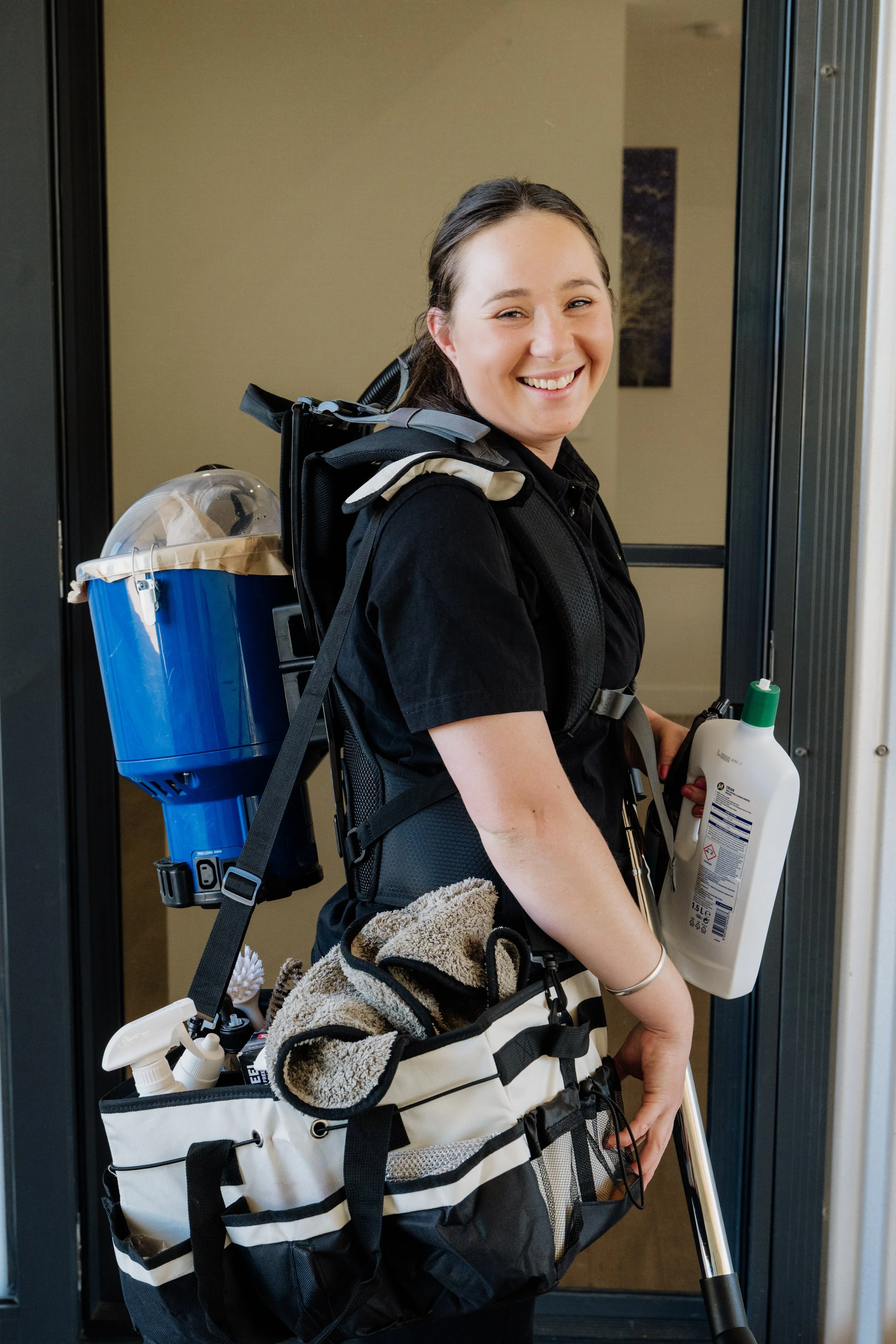A young woman with a backpack and a cart carrying cleaning supplies smiling in front of an elevator door.