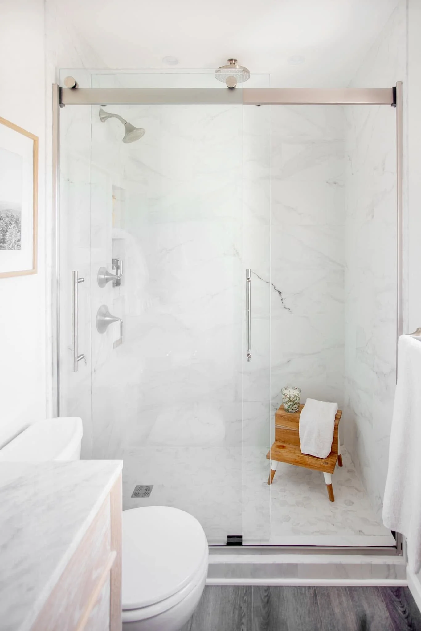 Bathroom with white marble walls and a glass shower enclosure. Inside the shower, there is a chrome showerhead and control panel, and a wooden stool with towels and a decorative jar. The bathroom has a paint or wallpaper artwork on the wall on the left side.