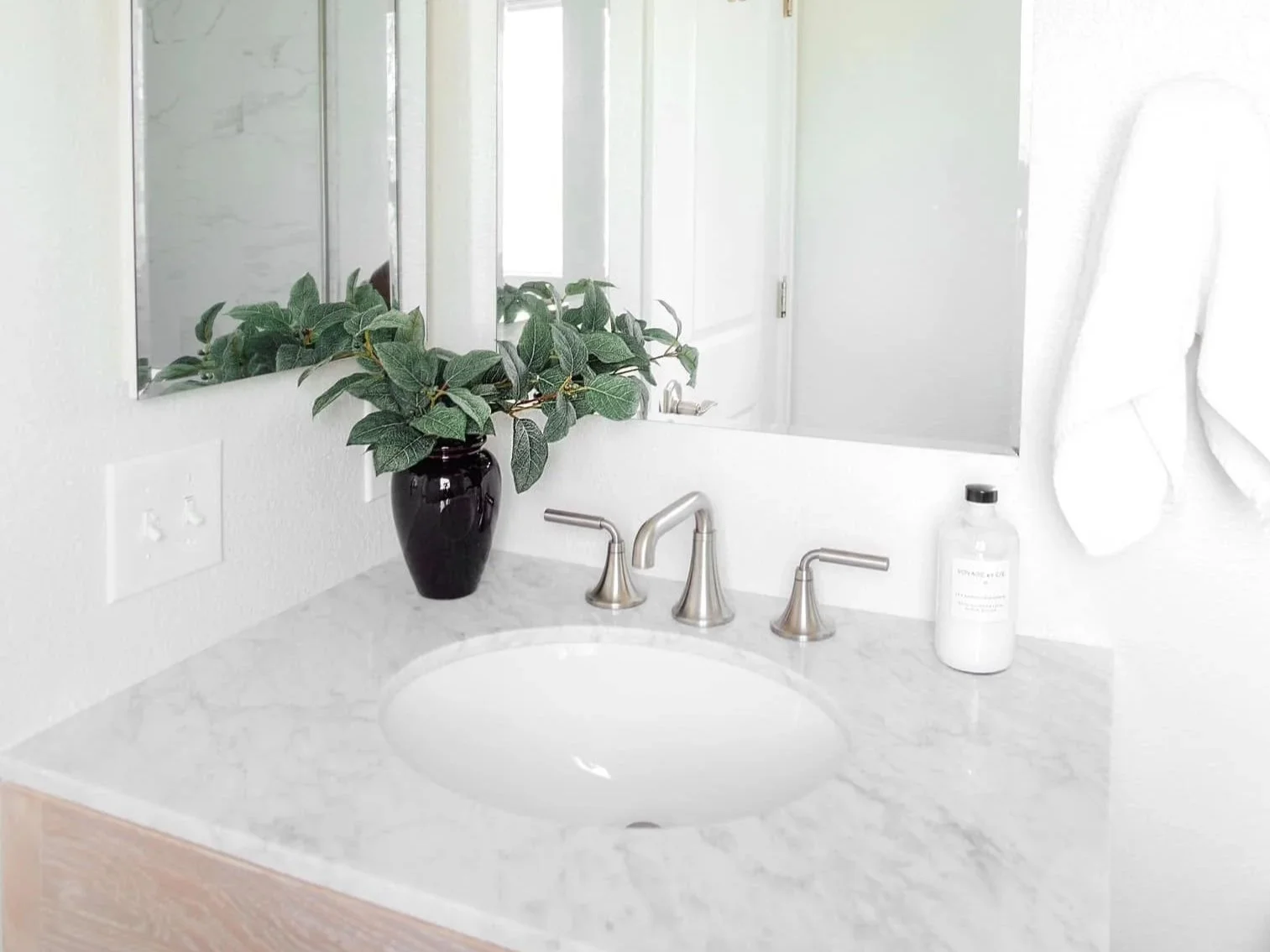 Bathroom vanity with a marble countertop, metal faucet fixtures, a black vase with green leaves, a bottle of liquid soap, and a white hand towel on a towel ring. Mirror reflects part of the room.
