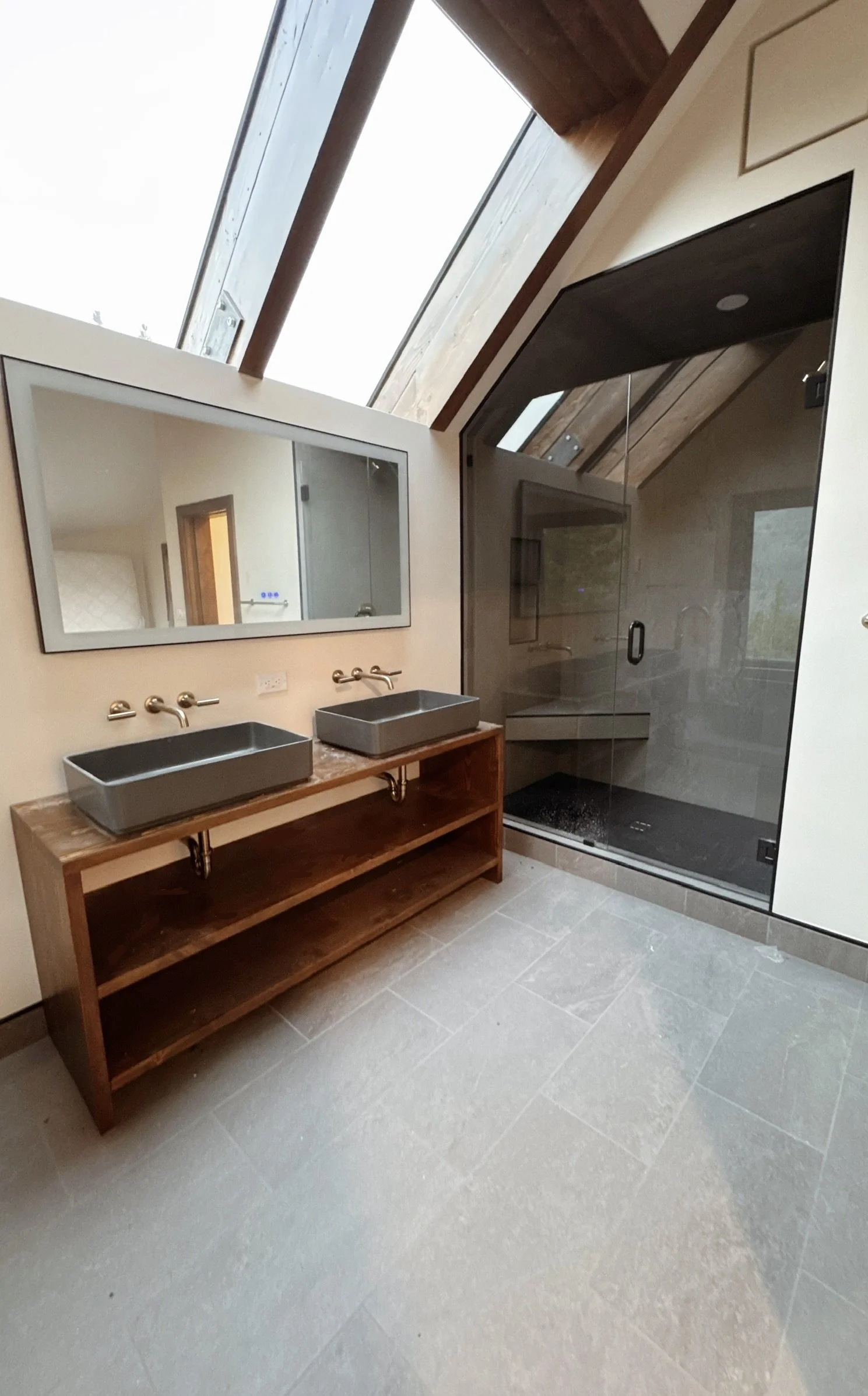 Bathroom with two rectangular vessel sinks on a wooden vanity, large mirror above, glass shower enclosure with dark trim, skylight with wooden framing, beige tile flooring.