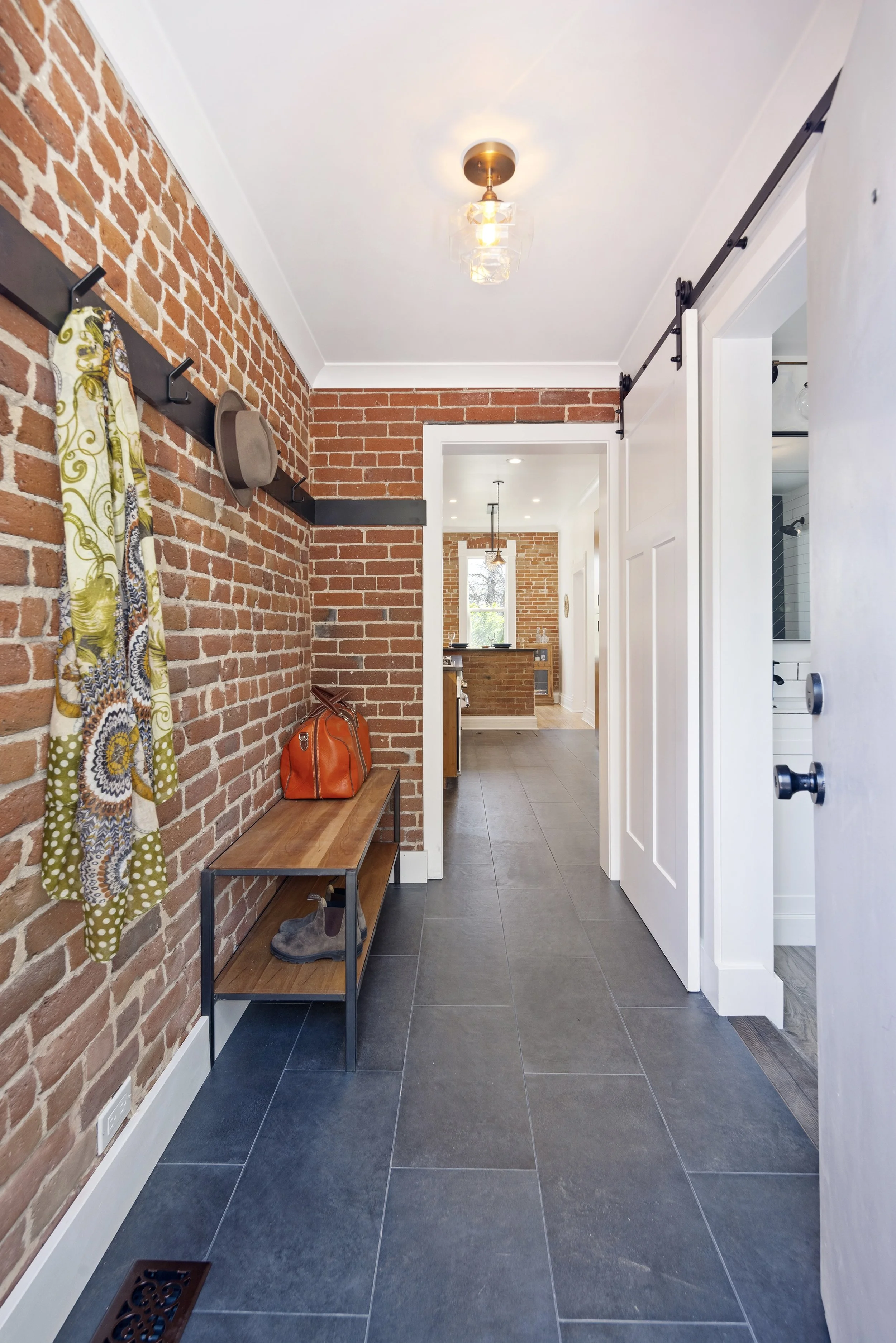 Entryway with exposed red brick wall, black hooks with coat, hat, and scarf, wooden bench with an orange bag on it, and dark gray tiled floor. Part of a white sliding barn door is visible on the right, and a doorway leads to a kitchen with more red brick walls and a window.