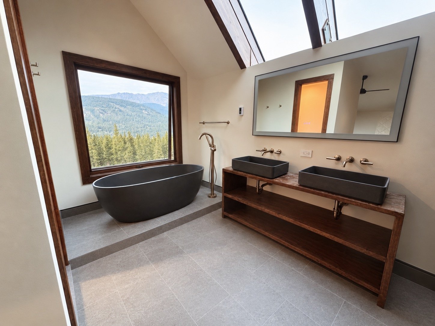Modern bathroom with black bathtub, large window with mountain view, dual black sinks on wooden vanity, large rectangular mirror, neutral-colored walls and floor, ceiling with skylights, and ceiling fan.