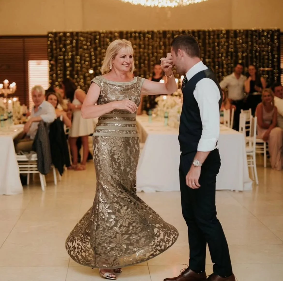 A woman in a gold, ornate evening dress dances with a man in a formal vest and white shirt at a wedding reception, with friends and family watching.