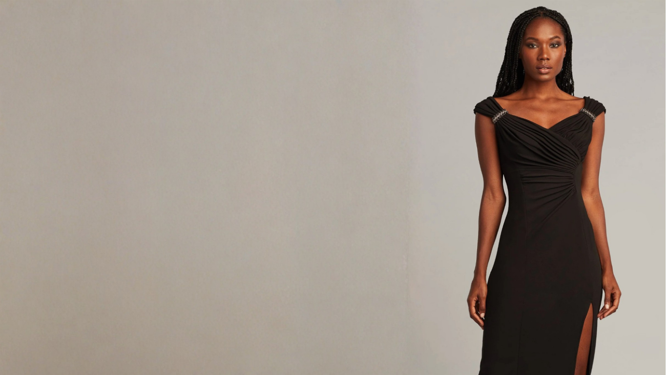 A woman with dark skin and long braids wearing a black evening dress with and slit on the side, standing against a plain light-colored background.