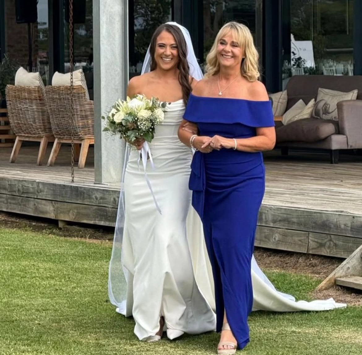 A bride in a white wedding dress holding a bouquet, standing next to a woman in a royal blue off-shoulder dress, at an outdoor wedding venue with a wooden deck and wicker chairs.