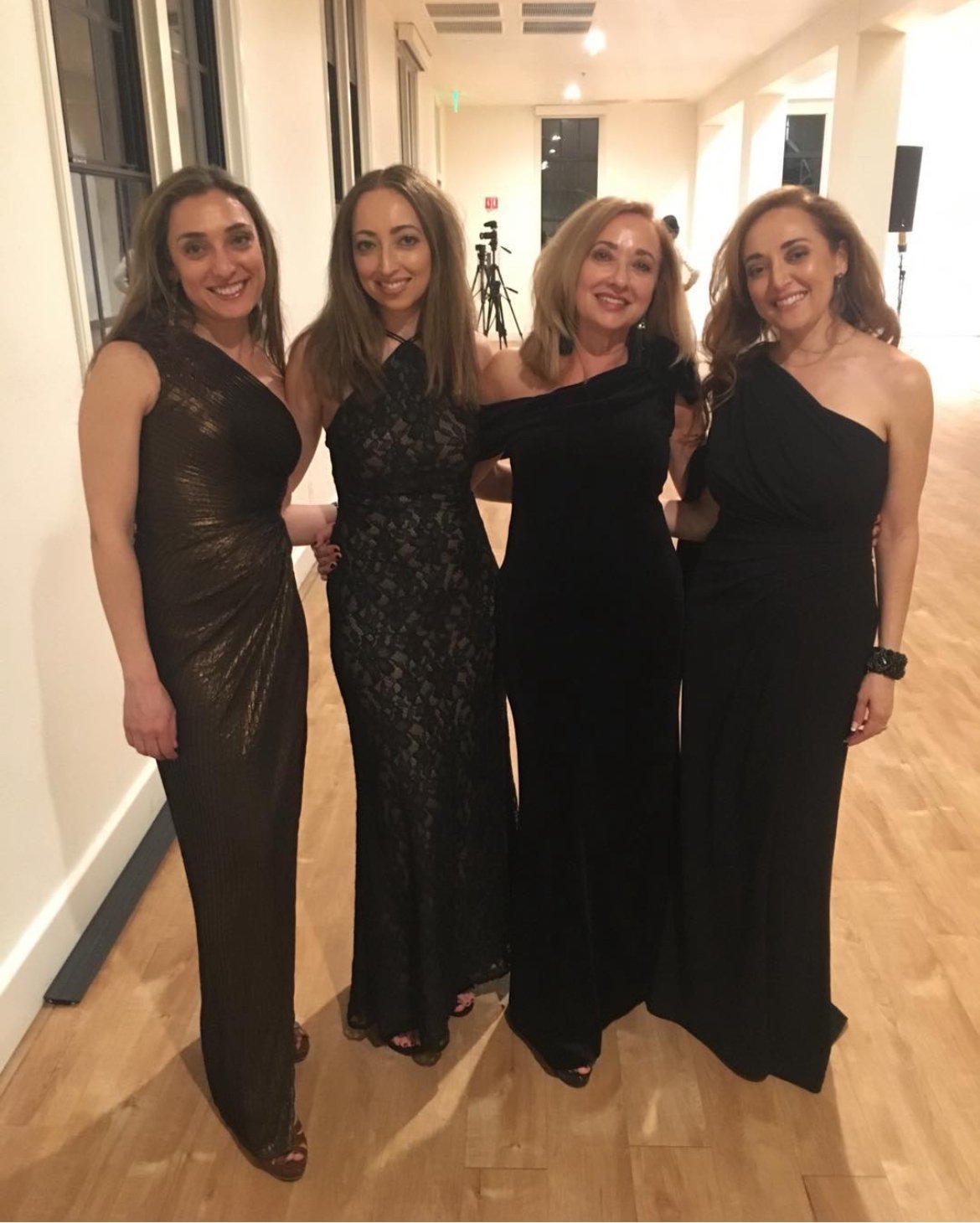 Four women dressed in black evening gowns standing together indoors, smiling for the camera at a social event.