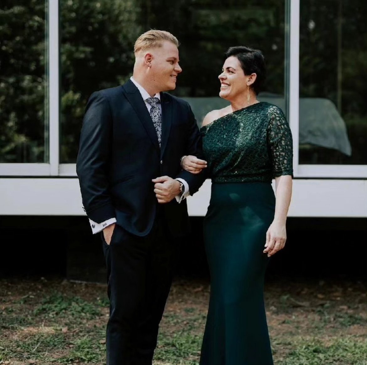 A young man in a dark suit and a woman in a dark green lace gown smiling while standing outdoors in front of a glass window.