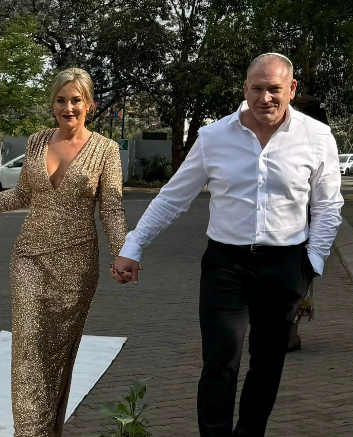 A couple holding hands and walking outdoors. The woman is wearing a gold, sequined, long-sleeved gown with a deep neckline. The man is dressed in a white shirt and black pants.