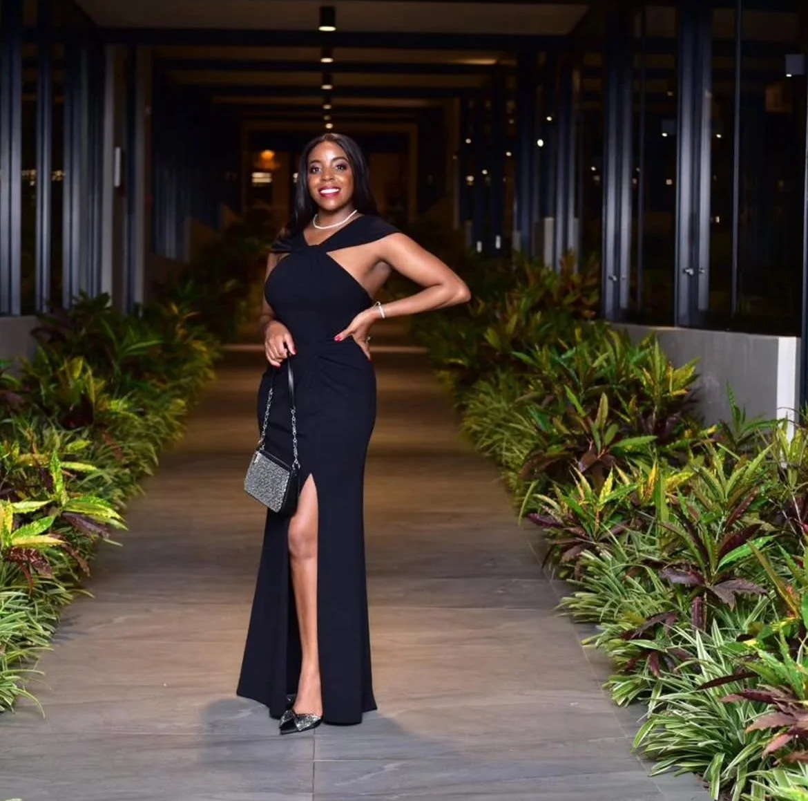A woman in a black evening gown with a thigh-high slit is standing on a wooden pathway at night, surrounded by lush plants. She is smiling, holding a glittery clutch, and wearing jewelry and heels.