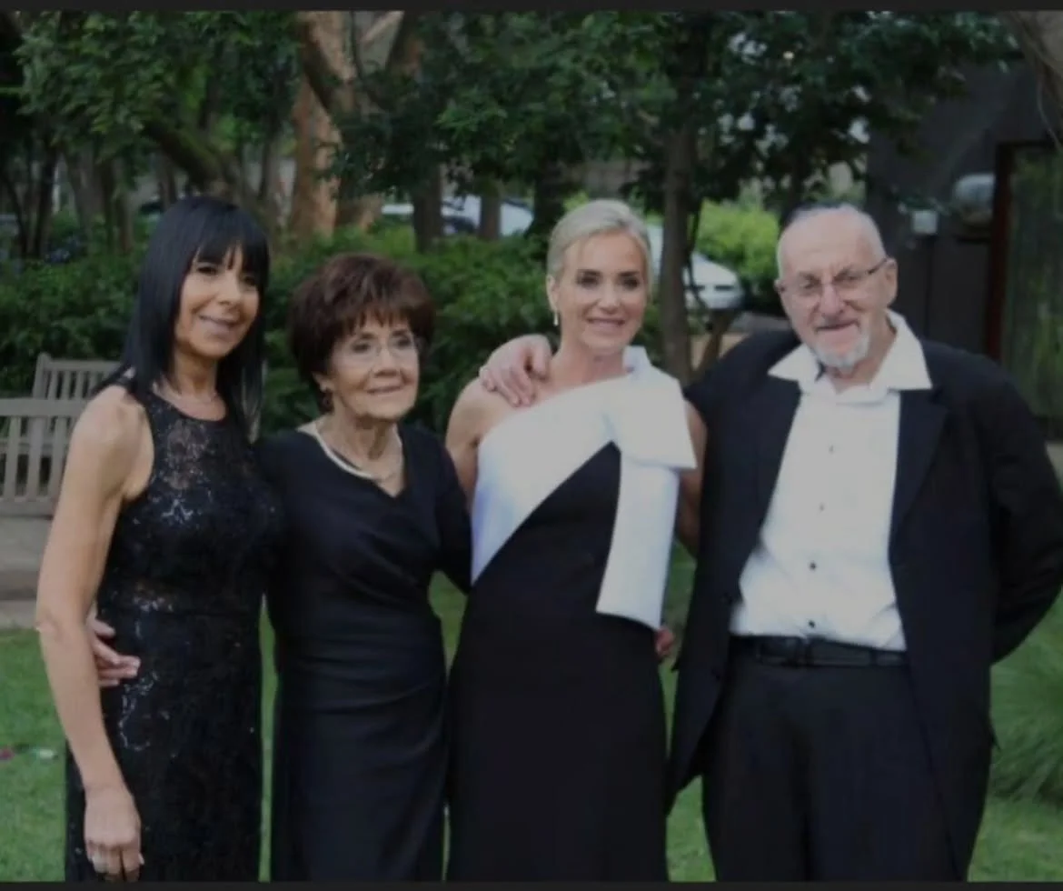 Four adults posing together outdoors, three women and one man, dressed in formal attire, smiling. They are standing on grass with trees and a park bench in the background.