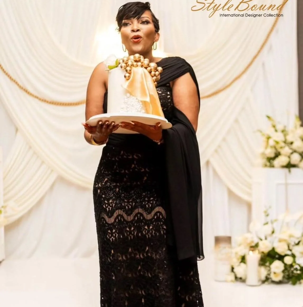 A woman in a black lace dress holding a decorative cake on a plate at a formal event with a cream-colored draped backdrop and flower arrangements in the background.