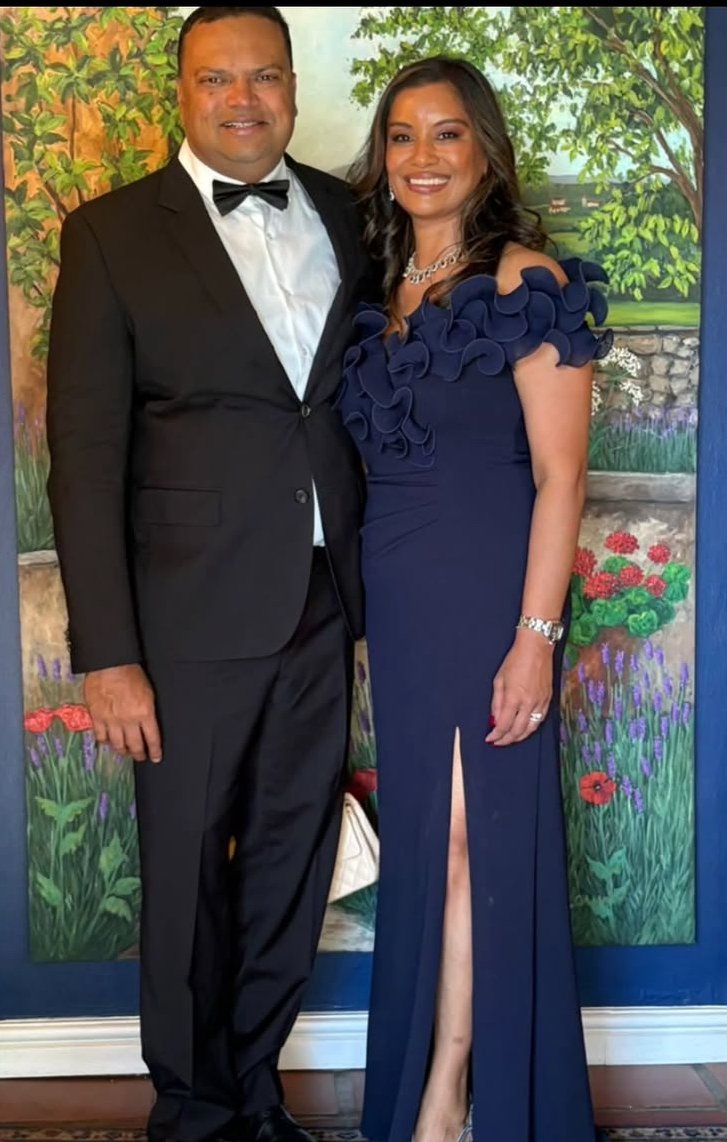 A man in a black tuxedo and bow tie standing next to a woman in an elegant navy blue gown with ruffle details and high slit, both smiling in front of a painted garden backdrop.