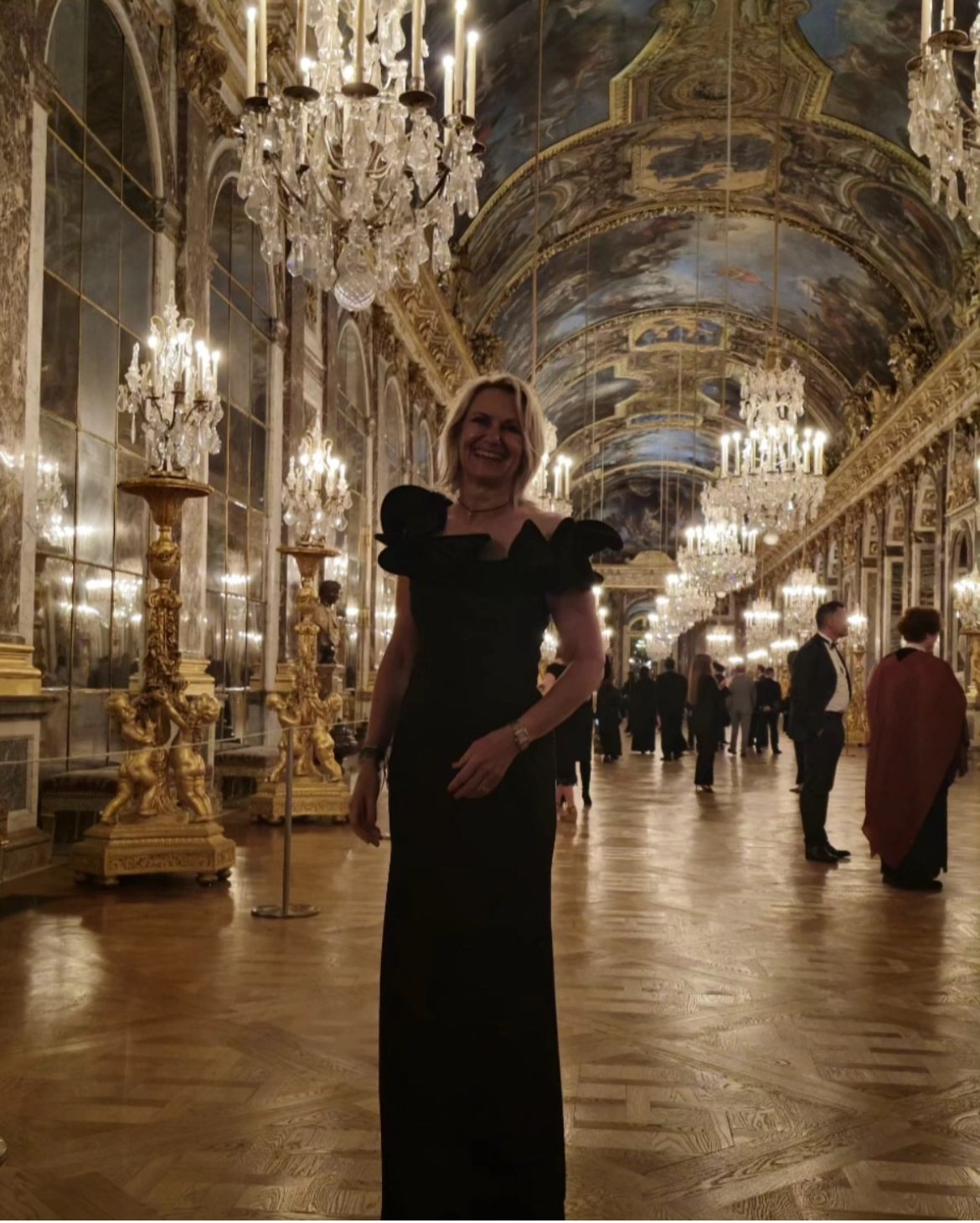 Woman in a black evening gown smiling in a grand, ornate hall with chandeliers, gilded decorations, and a painted ceiling.