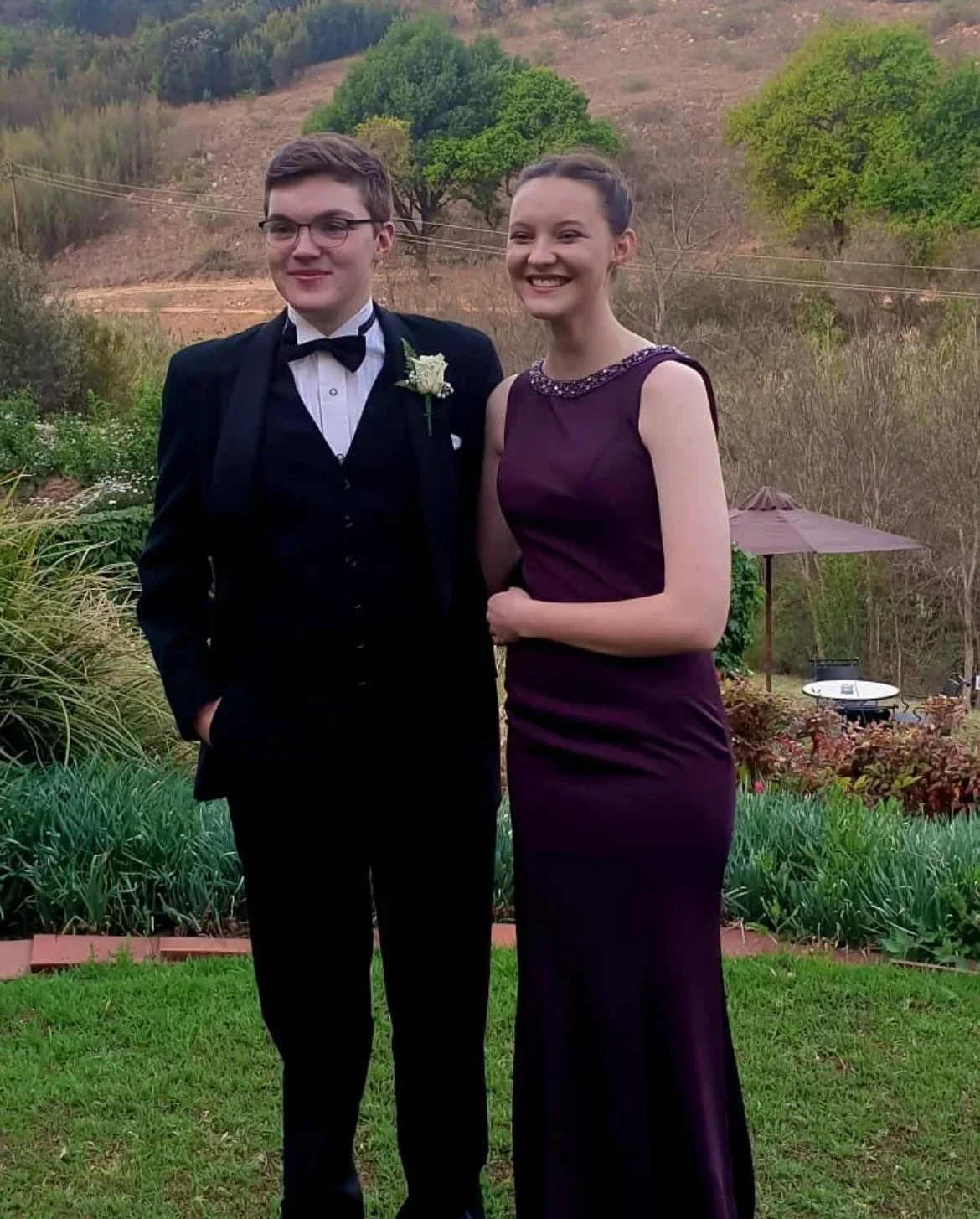 A young man in a tuxedo and a young woman in a purple evening gown standing outdoors in a garden with trees and a hillside in the background. They are smiling and posing for a photo.