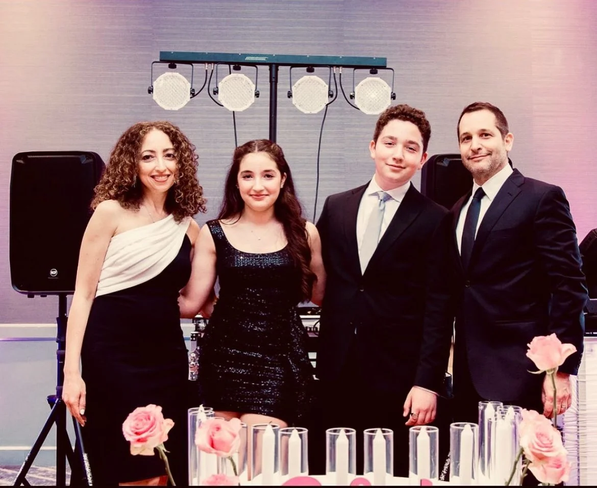 Group of four people dressed formally at a celebration event, standing behind a table with pink roses and candles, with a DJ setup in the background.