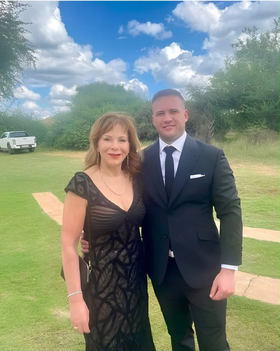 A woman and a man in formal attire posing outdoors on a sunny day, with a grassy area, trees, a dirt path, and a white vehicle in the background.