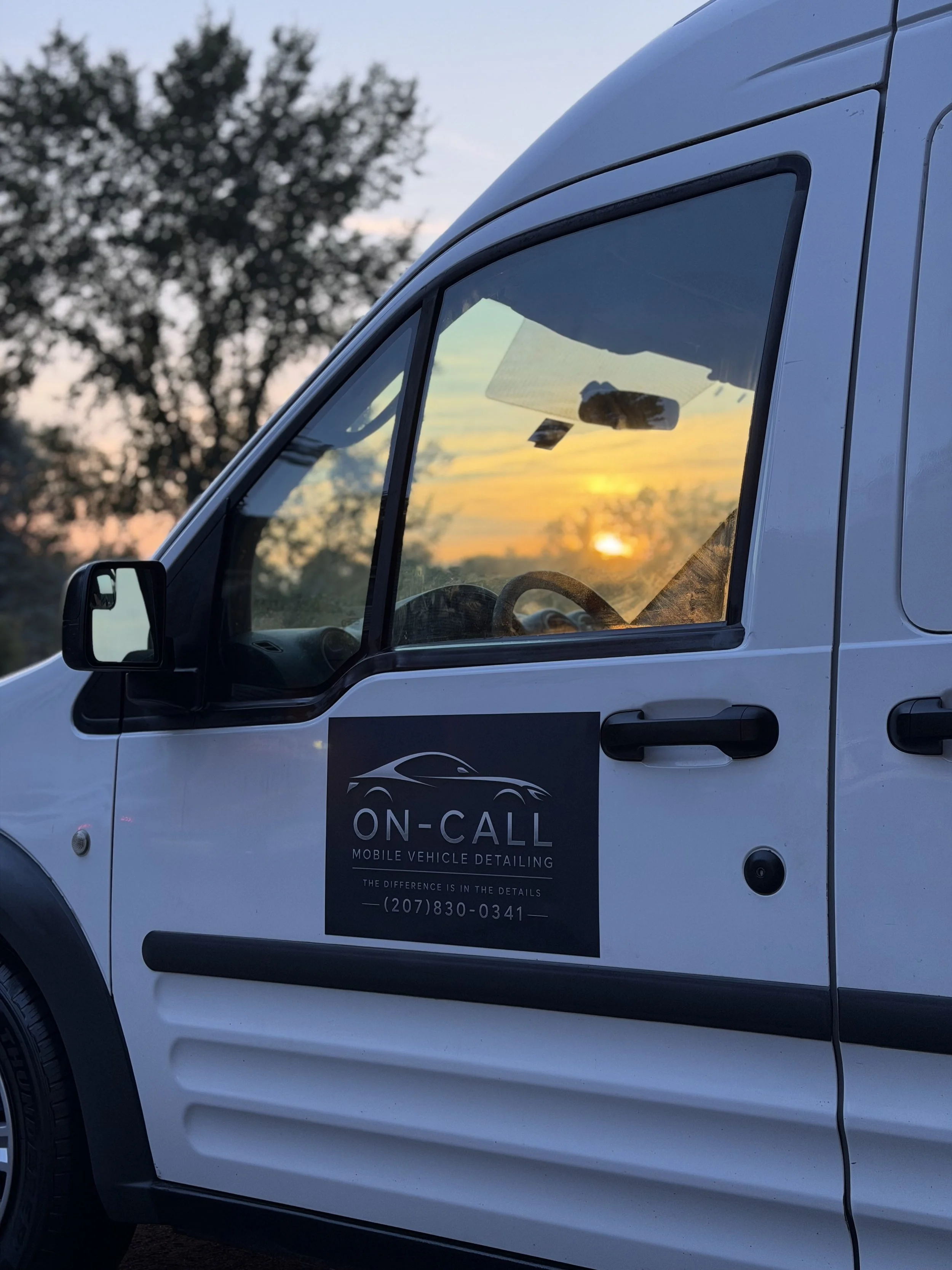 Close-up of a white vehicle with a sign on the door that reads "ON-CALL Mobile Vehicle Detailing." The background shows a sunset with trees and a cloudy sky reflected on the vehicle's window.