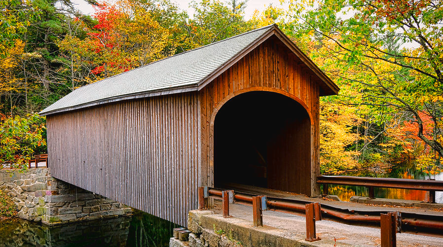 A wooden covered bridge over a small creek surrounded by autumn trees with colorful fall foliage.