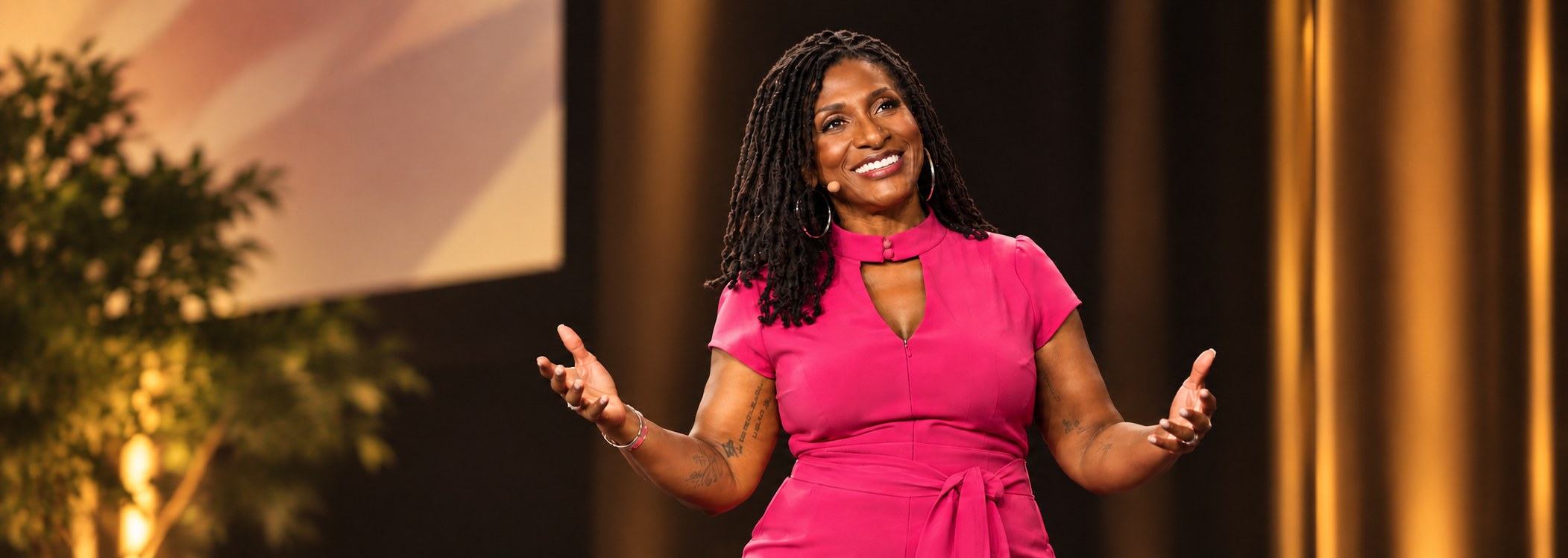 Dr. Amona Anderson, educator, veteran, and keynote speaker, with shoulder-length curly black hair, wearing a bright pink dress with a keyhole neckline, smiling and gesturing with her hands while speaking on stage.