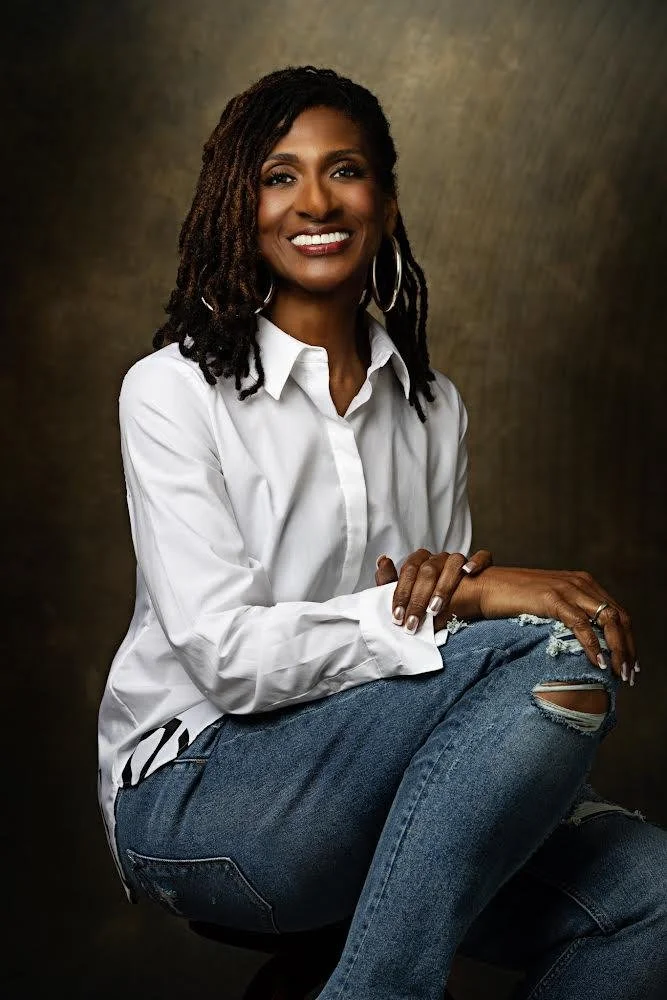 Dr. Amona Anderson, educator, veteran, and keynote speaker, a portrait of a Black woman with dreadlocks, wearing a white button-up shirt and ripped jeans, smiling against a dark, textured background.