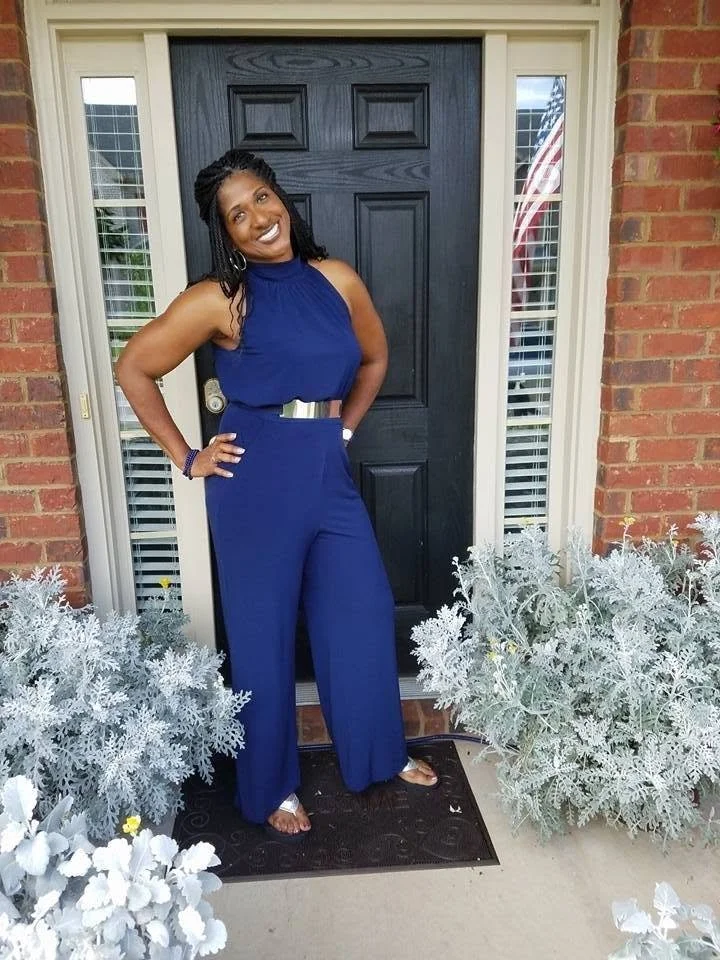 Dr. Amona Anderson, educator, veteran, and keynote speaker in a blue sleeveless jumpsuit standing at the front door of a house with brick walls, flanked by white plants, smiling.