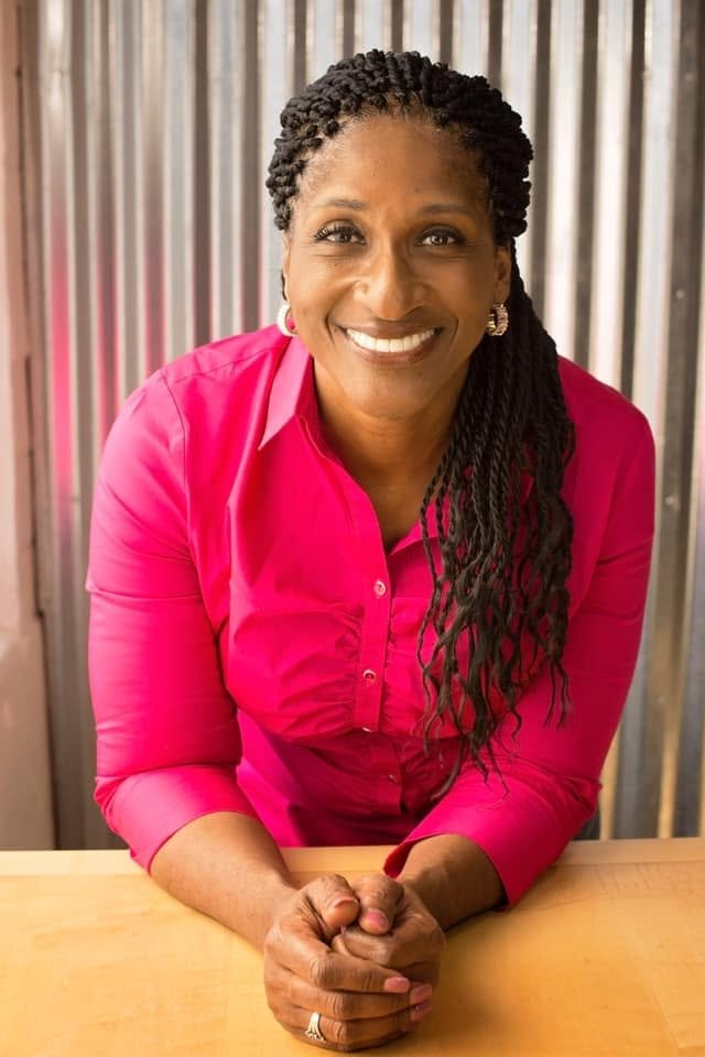 Dr. Amona Anderson, educator, veteran, and keynote speaker, smiling in a bright pink blouse sitting at a desk with her hands clasped, in front of a metallic slat background.