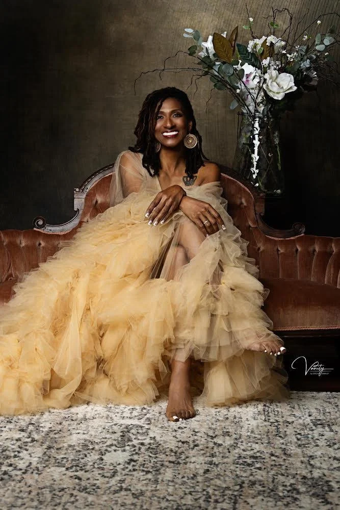 Dr. Amona Anderson, educator, veteran, and keynote speaker with dark skin, sitting on a vintage brown velvet sofa, wearing a beige tulle gown. Behind her is a large floral arrangement with white flowers and greenery, in a tall glass vase.