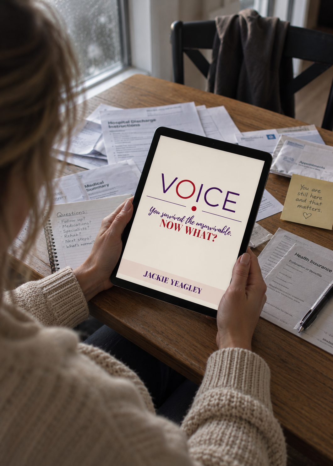 Woman sitting at a wooden table holding a tablet with the book title 'Voice: You survived the unthinkable. Now what?' by Jackie Yeagley, surrounded by medical documents, notes, and papers.